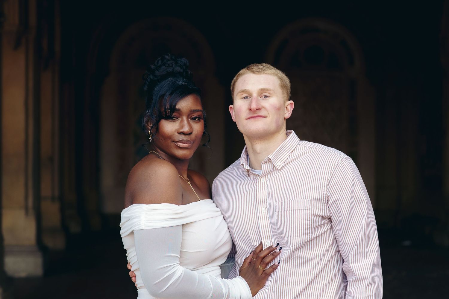A couple poses together outdoors, with one wearing a white off-shoulder dress and the other in a light pink button-up shirt.