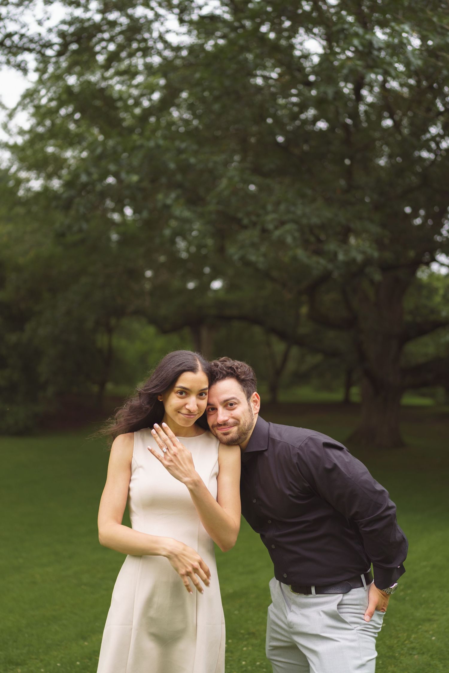 A couple stands close together smiling on a lush green lawn with trees in the background.