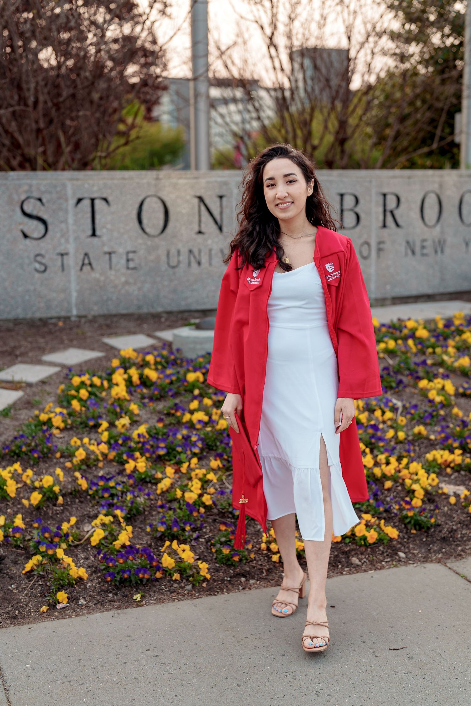 A person in a red graduation gown poses in front of the Stony Brook University sign with yellow flowers blooming nearby.