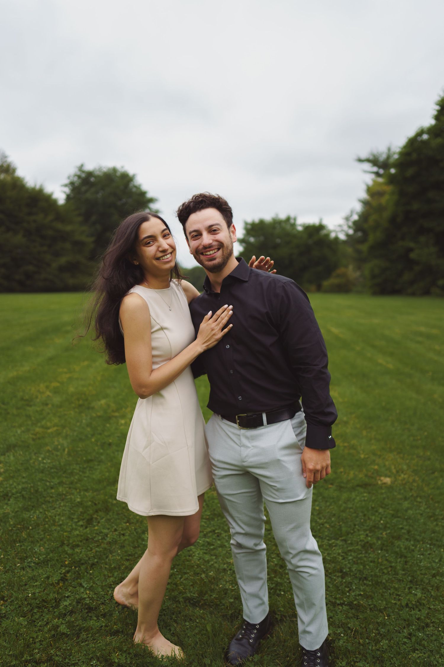 A couple laughs together while posing playfully in a green field on an overcast day.