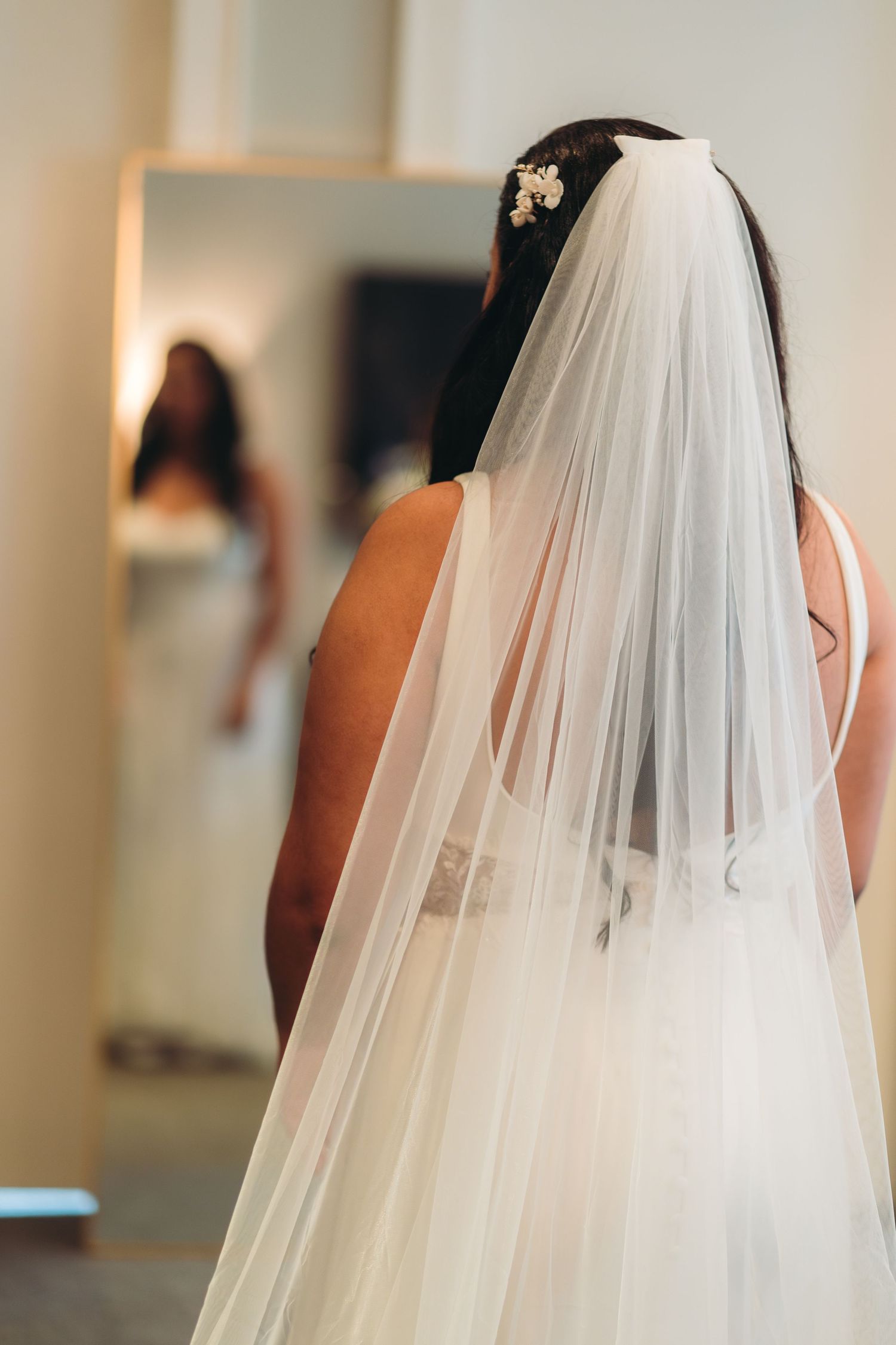 Back view of a bride wearing an elegant flowing white veil looking at her reflection in a dressing room mirror.