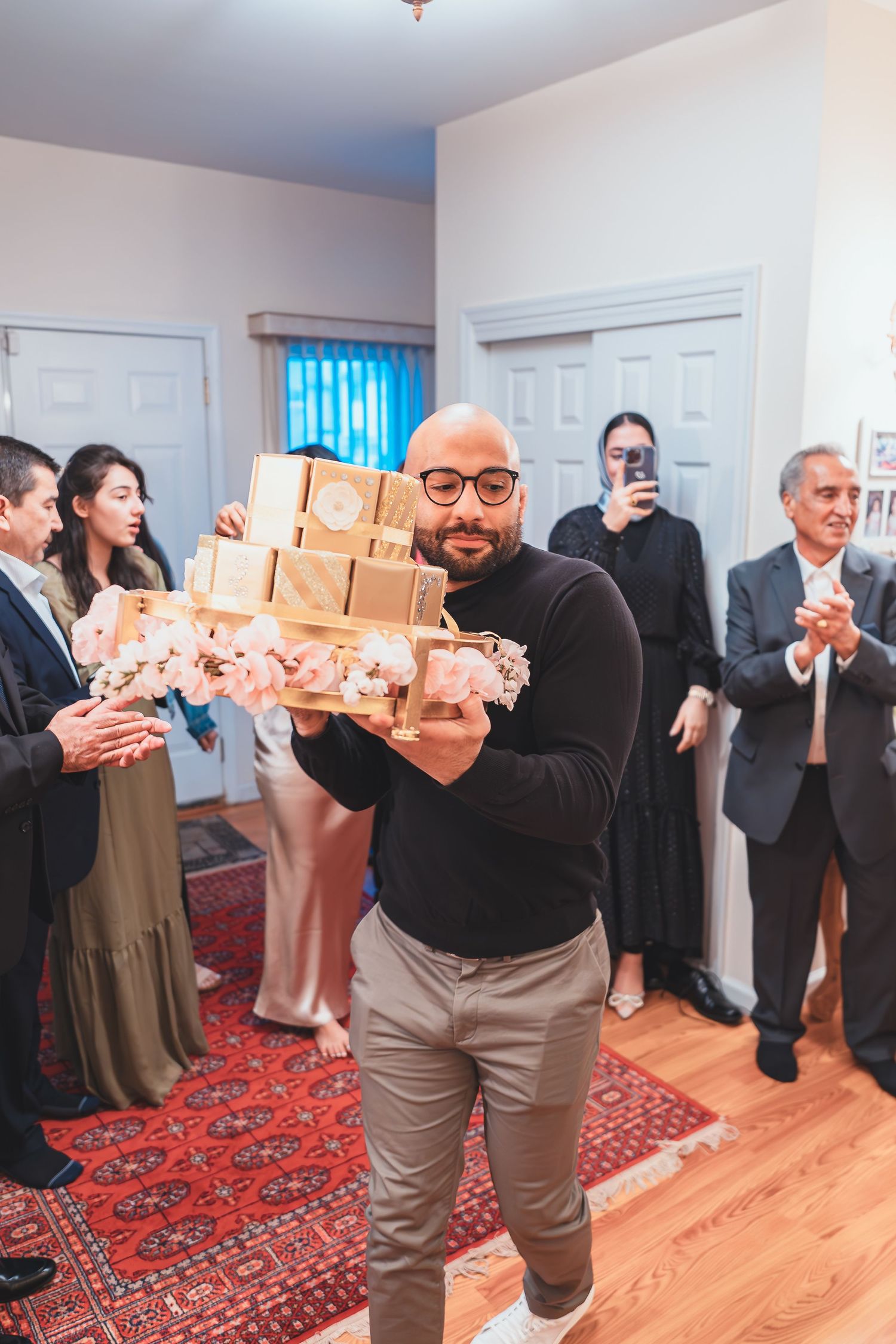 A person in black holds stacked gift boxes while guests applaud in a room with hardwood floors.