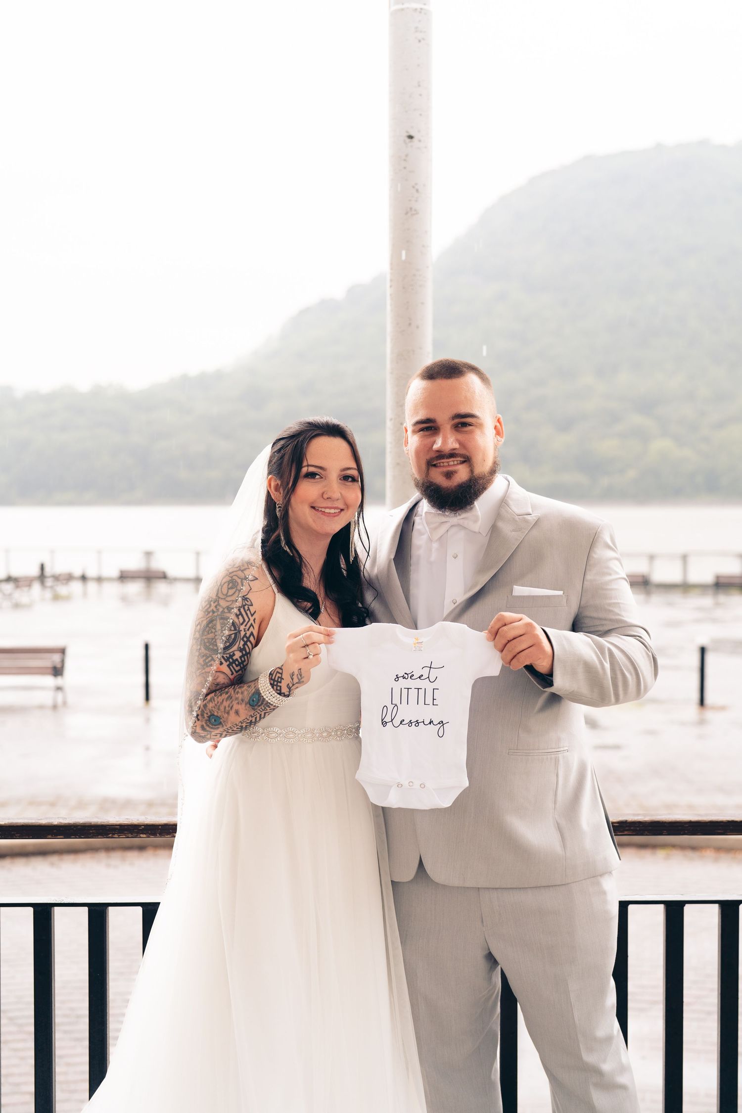 A couple in wedding attire holds up a small white onesie on a lakeside deck.