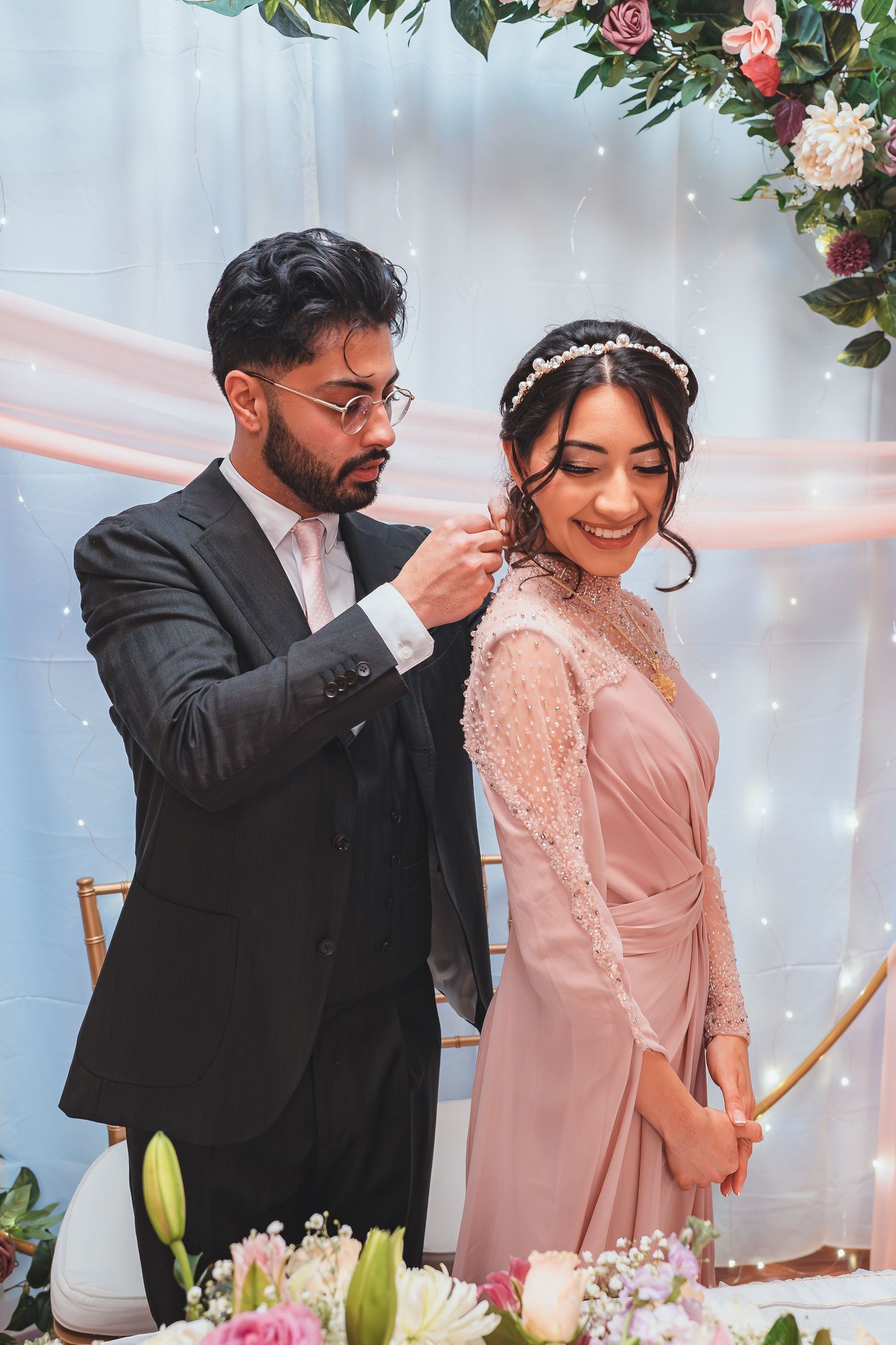 A couple in pink and black formal wear shares a joyful moment by floral decorations and twinkling lights.