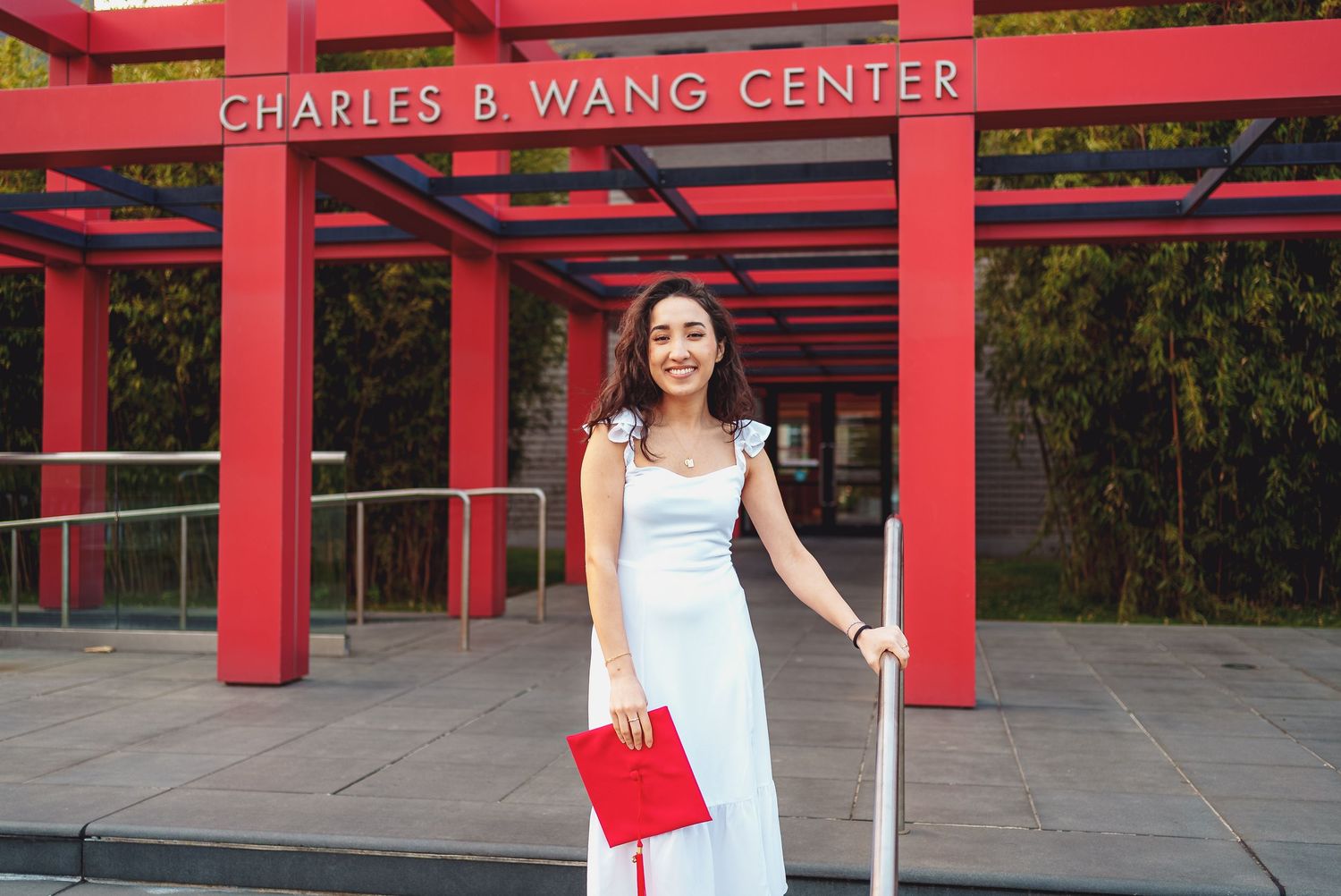 Visitor in white dress stands outside Charles B. Wang Center entrance with red beams and architectural details.