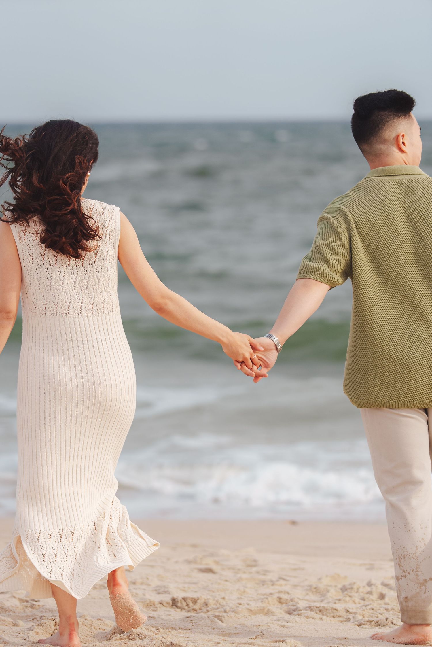 Couple holding hands and walking on beach with woman in white dress and man in tan pants and olive shirt.