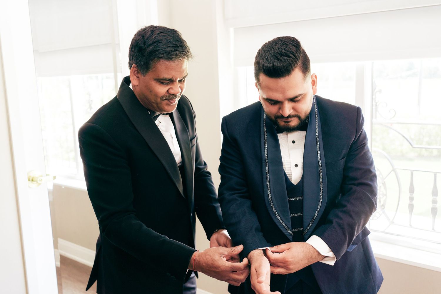 Two people wearing formal tuxedos help each other prepare before a special event.