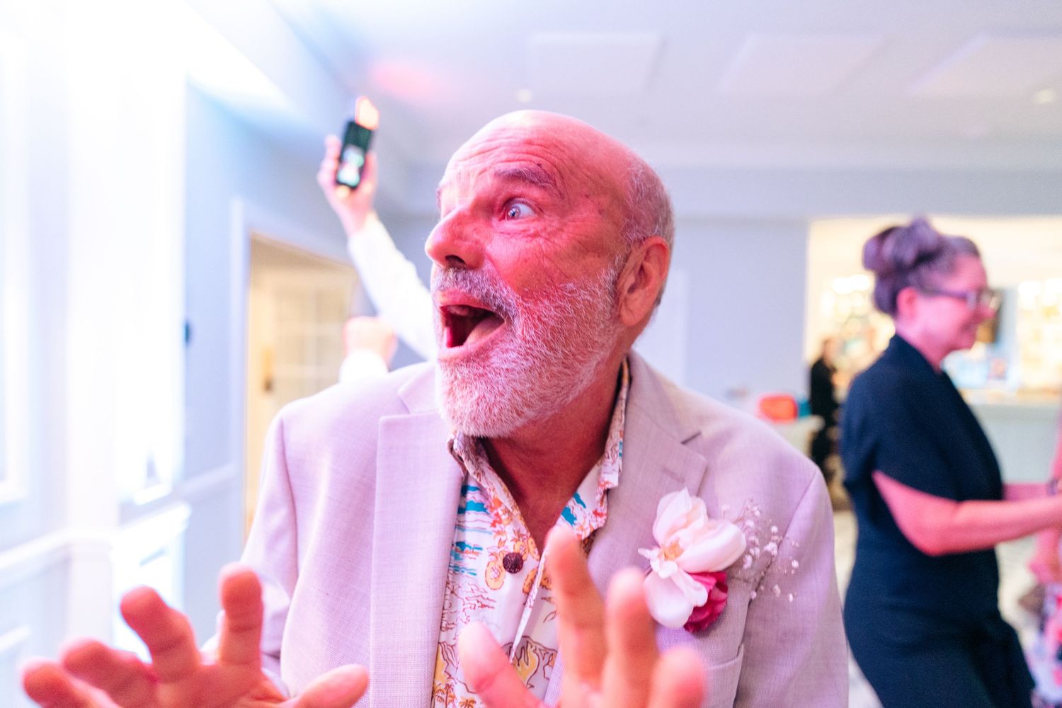 Energetic person in a light pink blazer and floral boutonniere gestures animatedly while holding a phone at an indoor event.