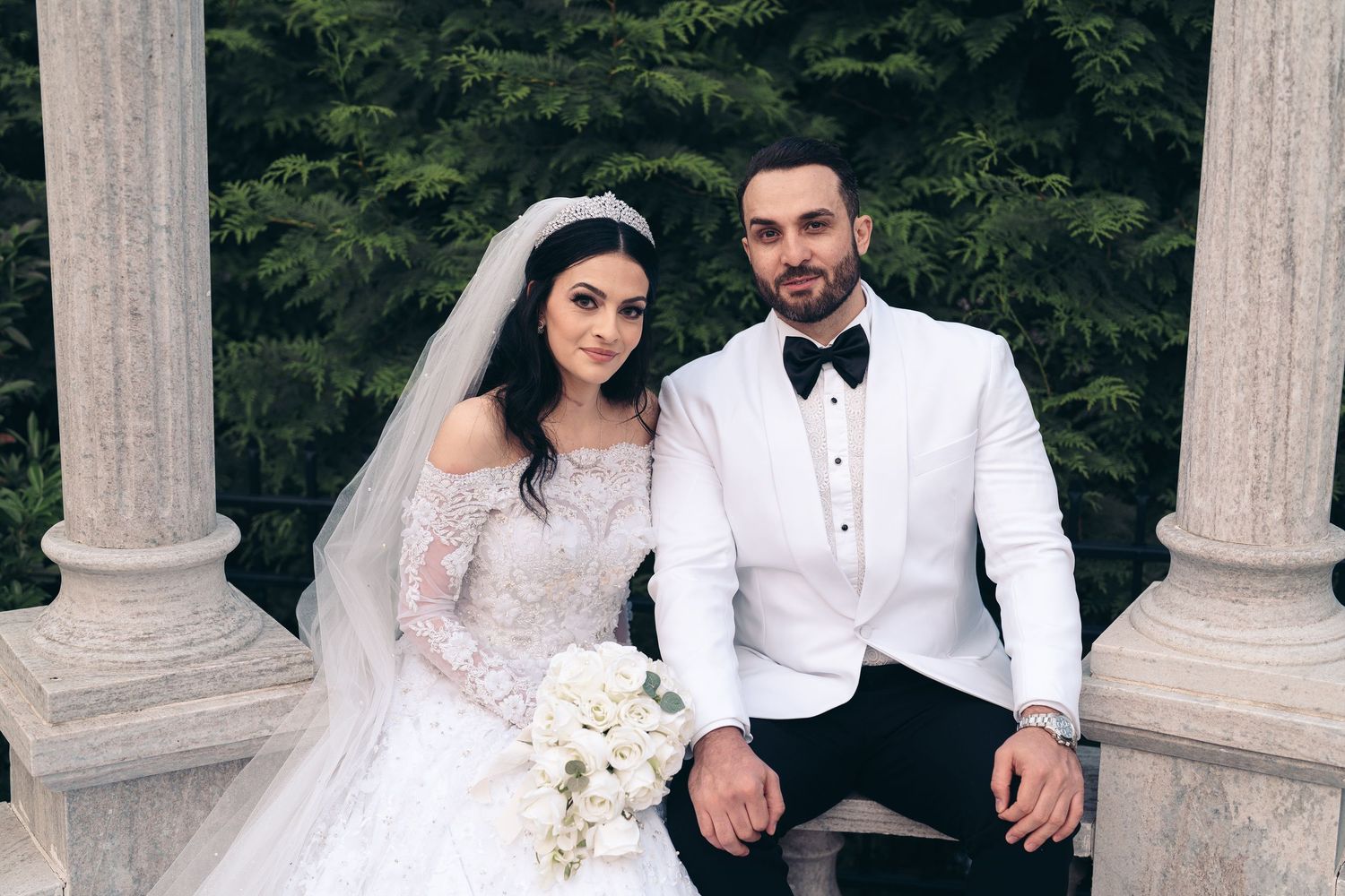 A bride in a lace wedding gown and groom in white tuxedo sit between stone pillars holding a white floral bouquet.