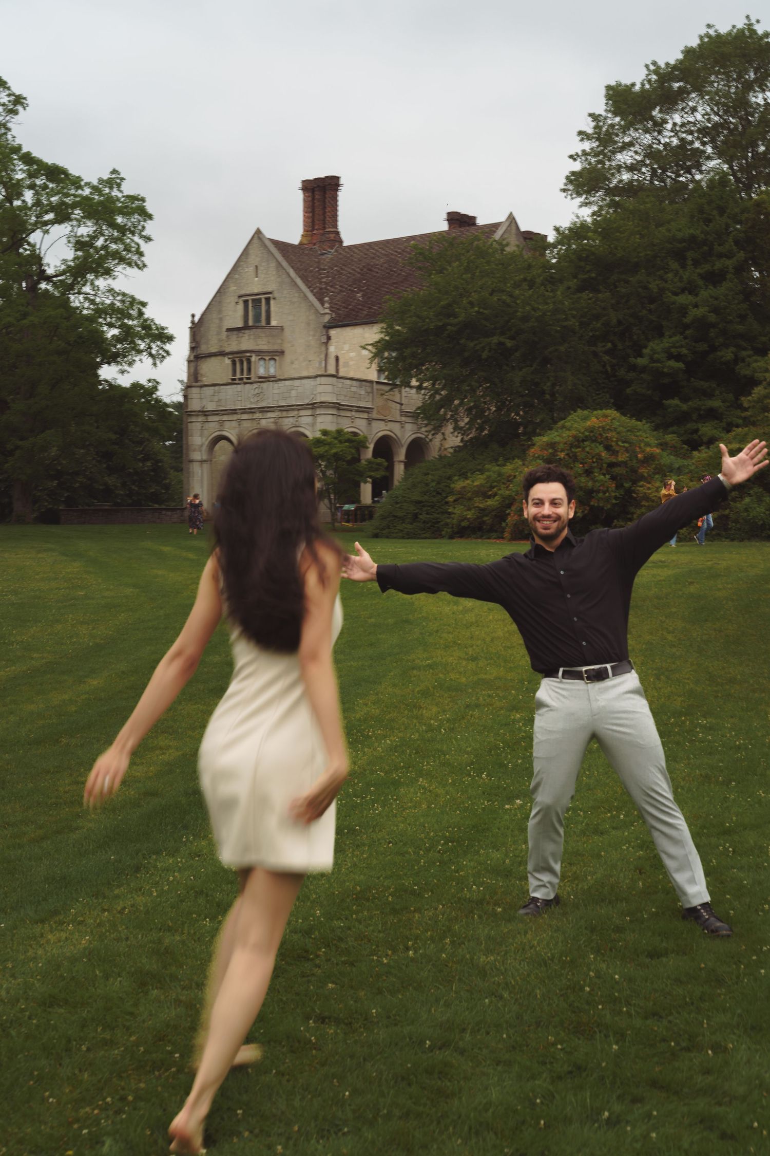A couple celebrating with joy on a lawn in front of a stately stone mansion with arms outstretched.