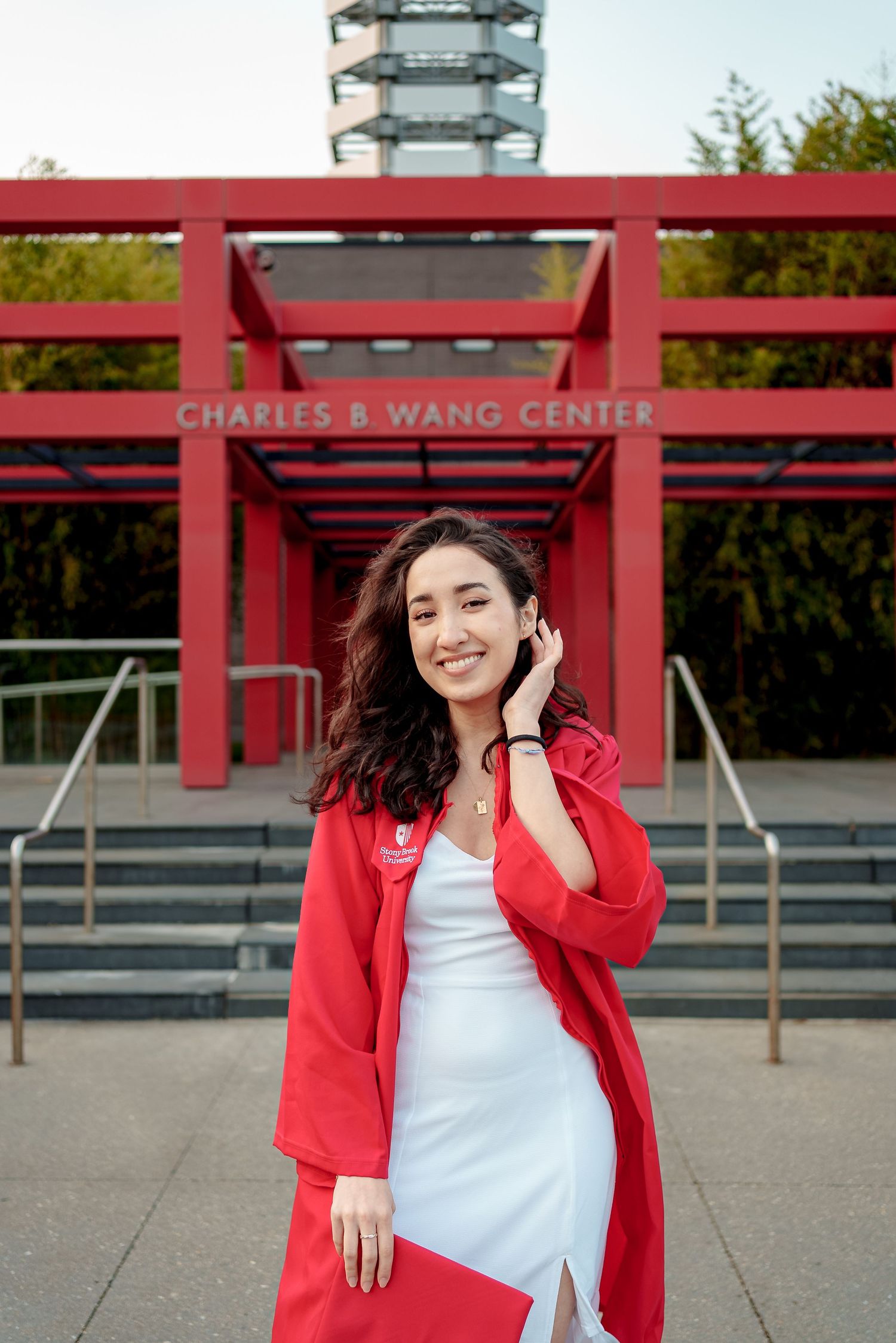 Student wearing a red coat and white dress poses outside the Charles B. Wang Center on a sunny day.