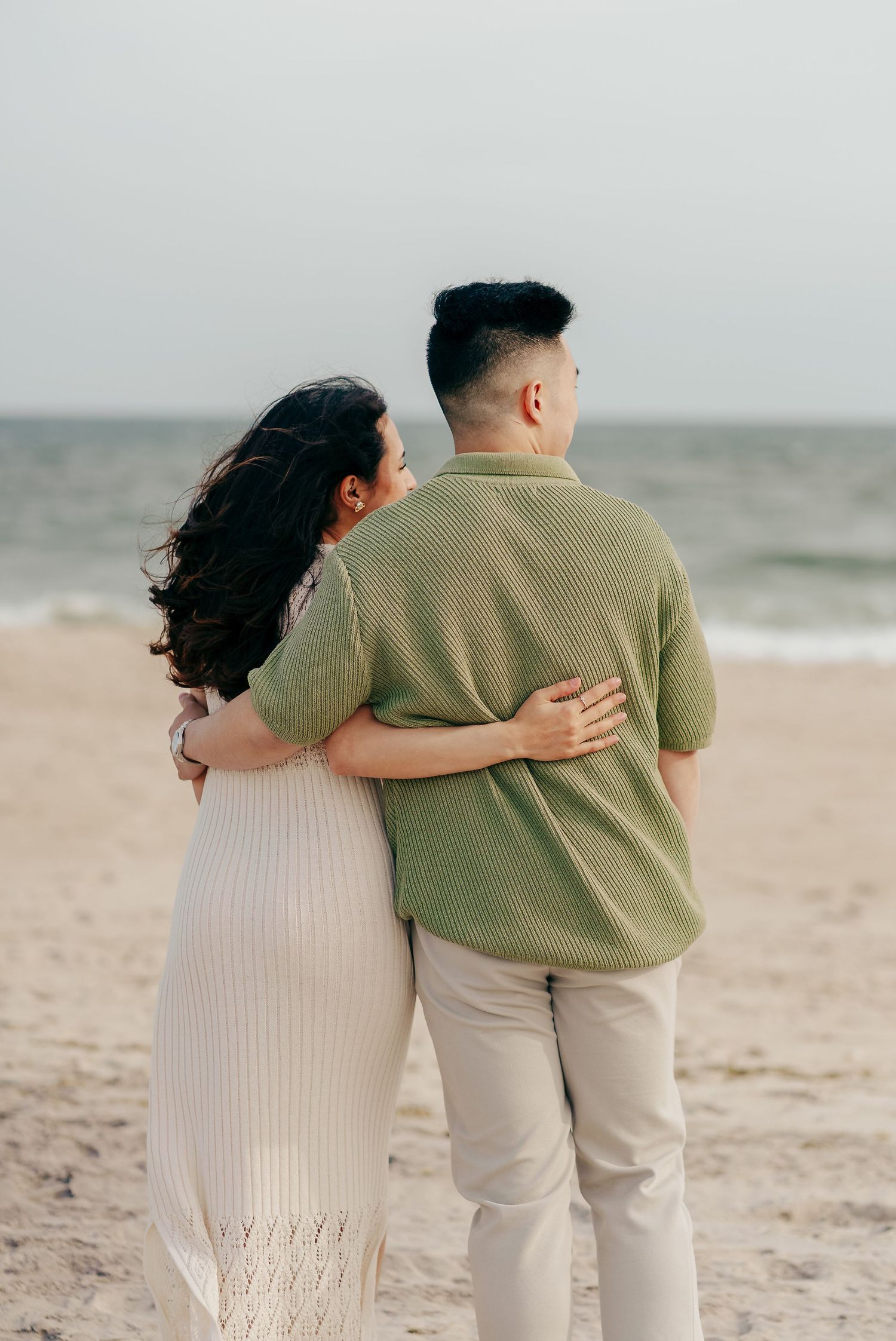 A romantic couple embraces while looking out at a misty beach, wearing beige and green casual attire.