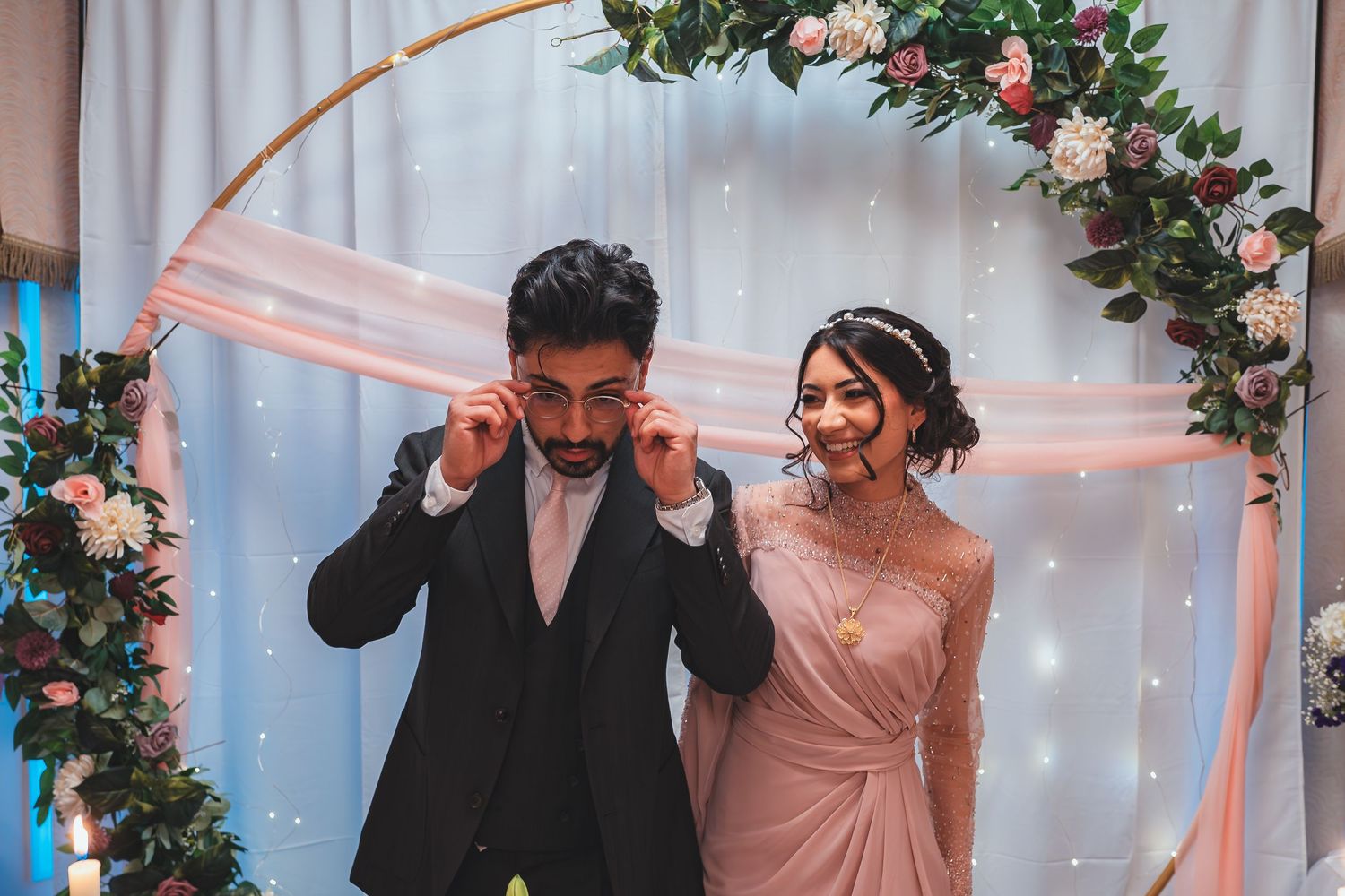 A couple shares a playful moment under an arch decorated with pink roses and fairy lights.
