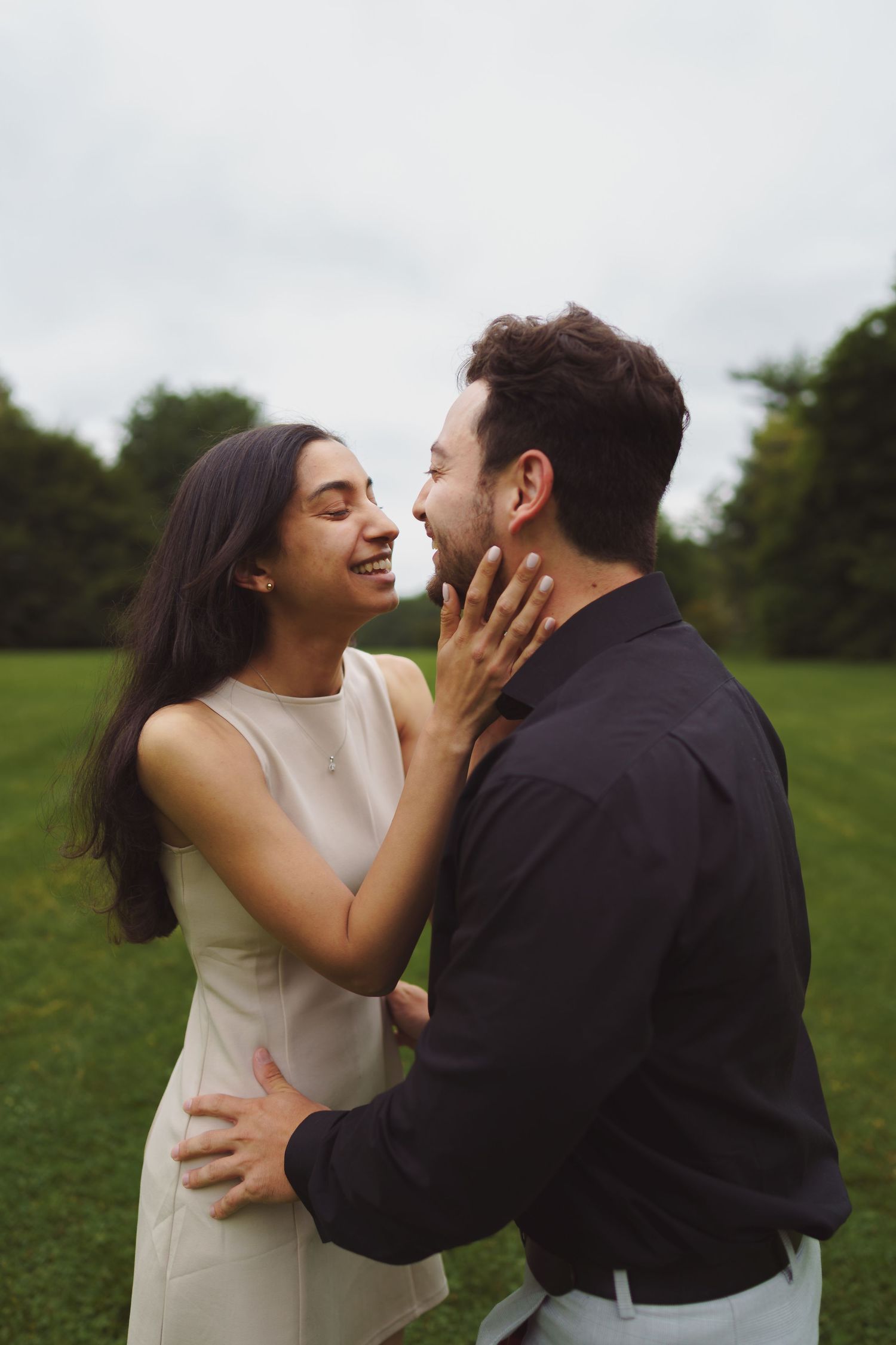 A couple in formal attire playfully interacts in a green outdoor field during a romantic photoshoot.