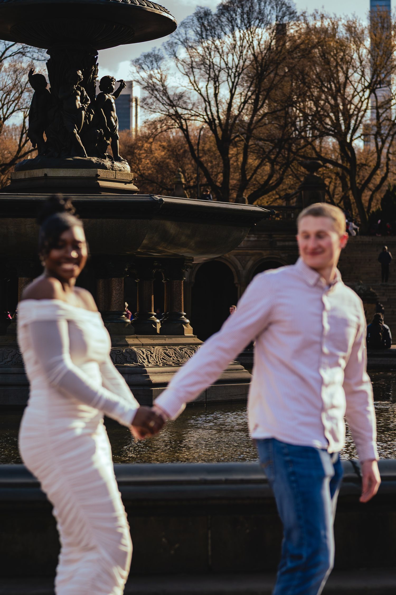 A couple in contrasting white and blue outfits holds hands near an ornate fountain in Central Park during autumn.
