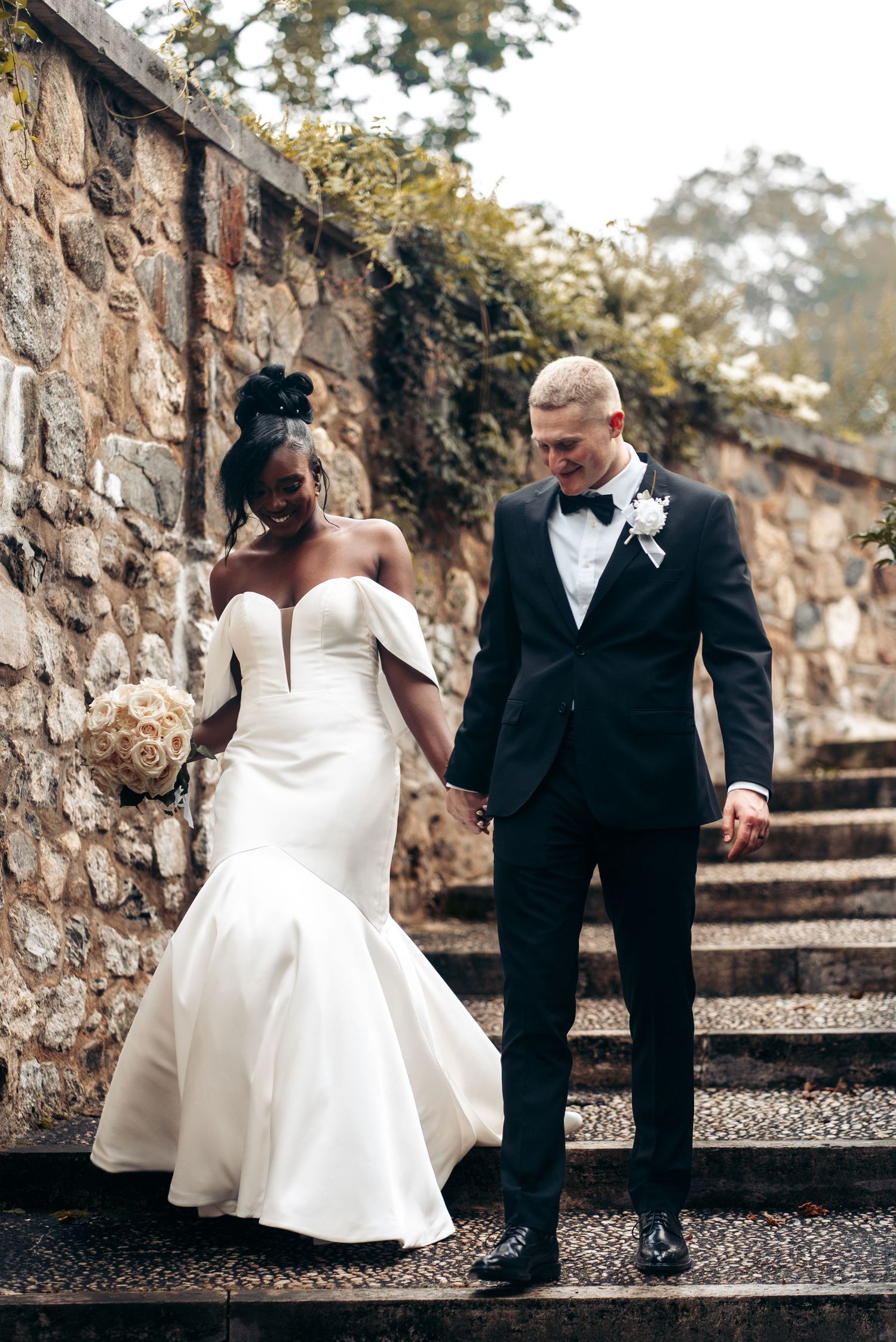 A couple in wedding attire walks down stone steps against a rustic stone wall backdrop outdoors.