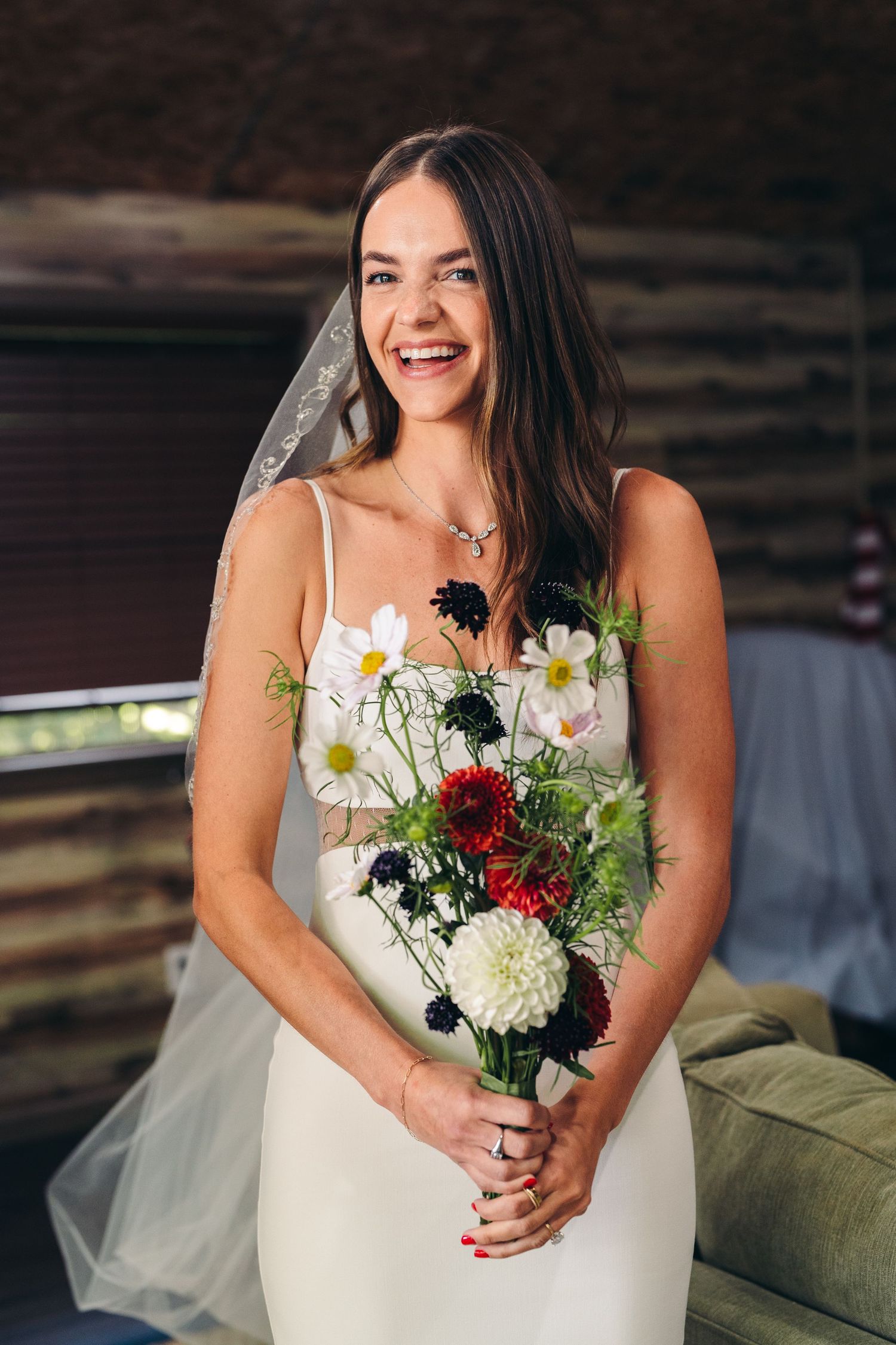 A happy bride wearing a white spaghetti strap wedding dress holds a vibrant bouquet with red and white flowers.