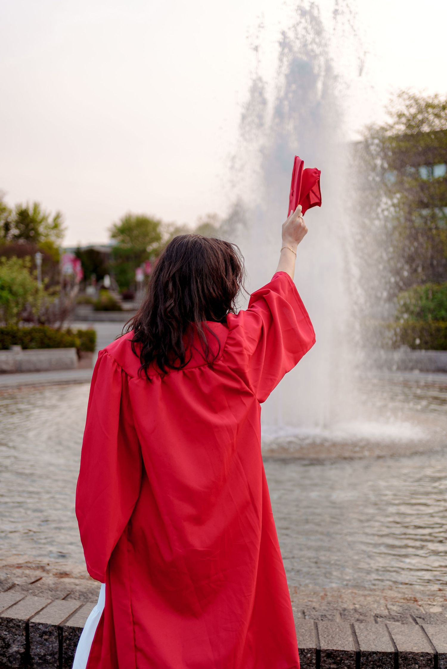 Graduate in red cap and gown raises diploma in front of outdoor fountain during commencement celebration.