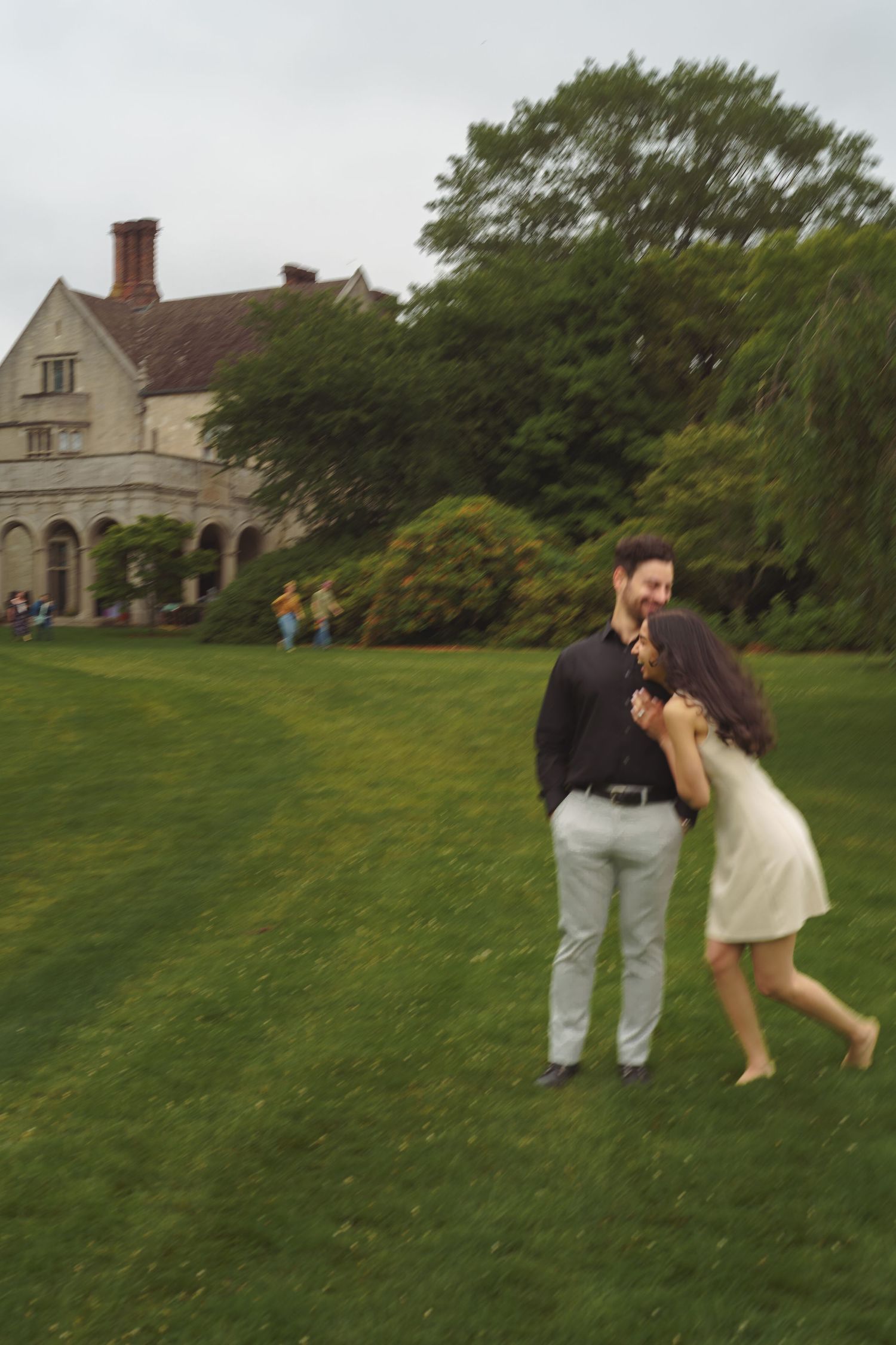 Two people share a romantic moment on a grassy lawn in front of a stone mansion on a summer day.