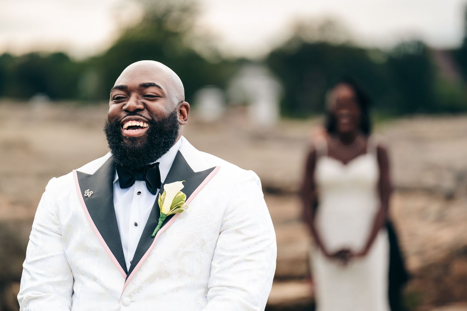 A joyful groom wearing a white tuxedo jacket and black bowtie laughs heartily during an outdoor wedding photoshoot.