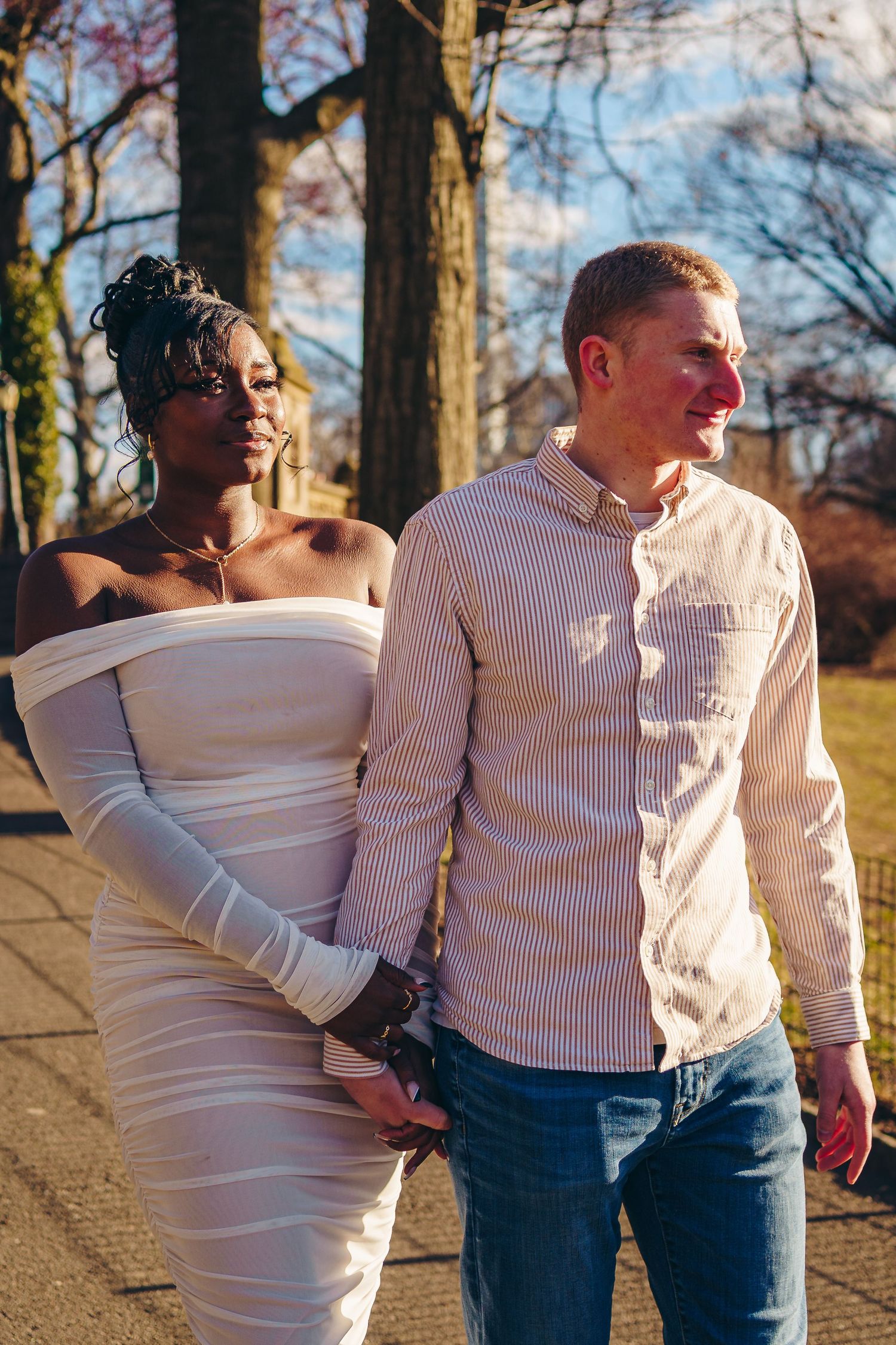 Two happy people share a warm embrace while walking outdoors in the sunlight along a tree-lined path.