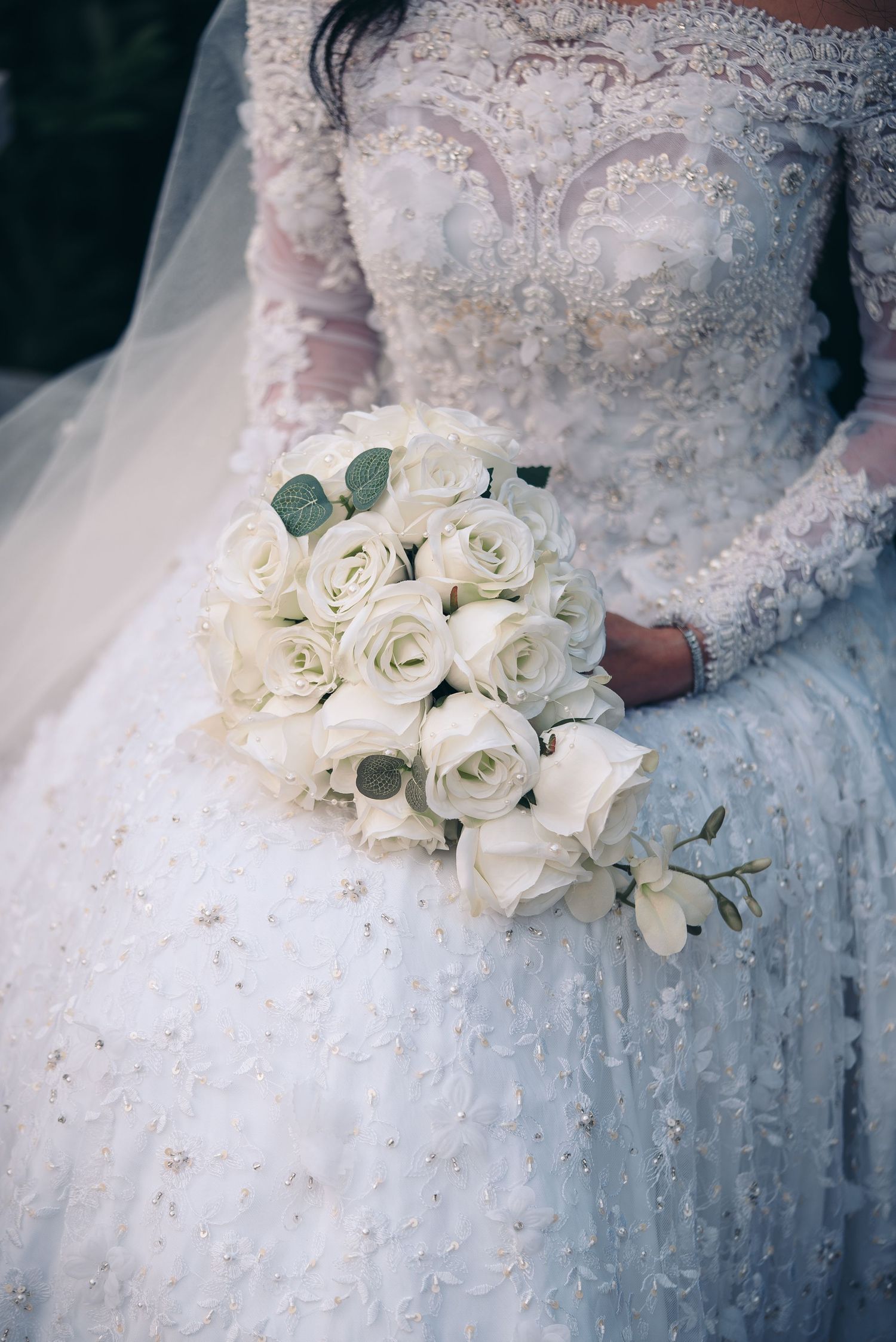 A classic white rose wedding bouquet held against an ornate lace wedding dress with delicate beading details.