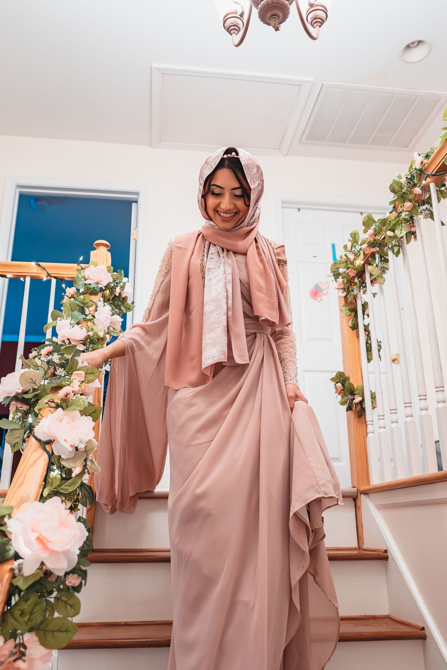 A woman in a dusty pink hijab and matching dress stands elegantly on a staircase decorated with floral arrangements.