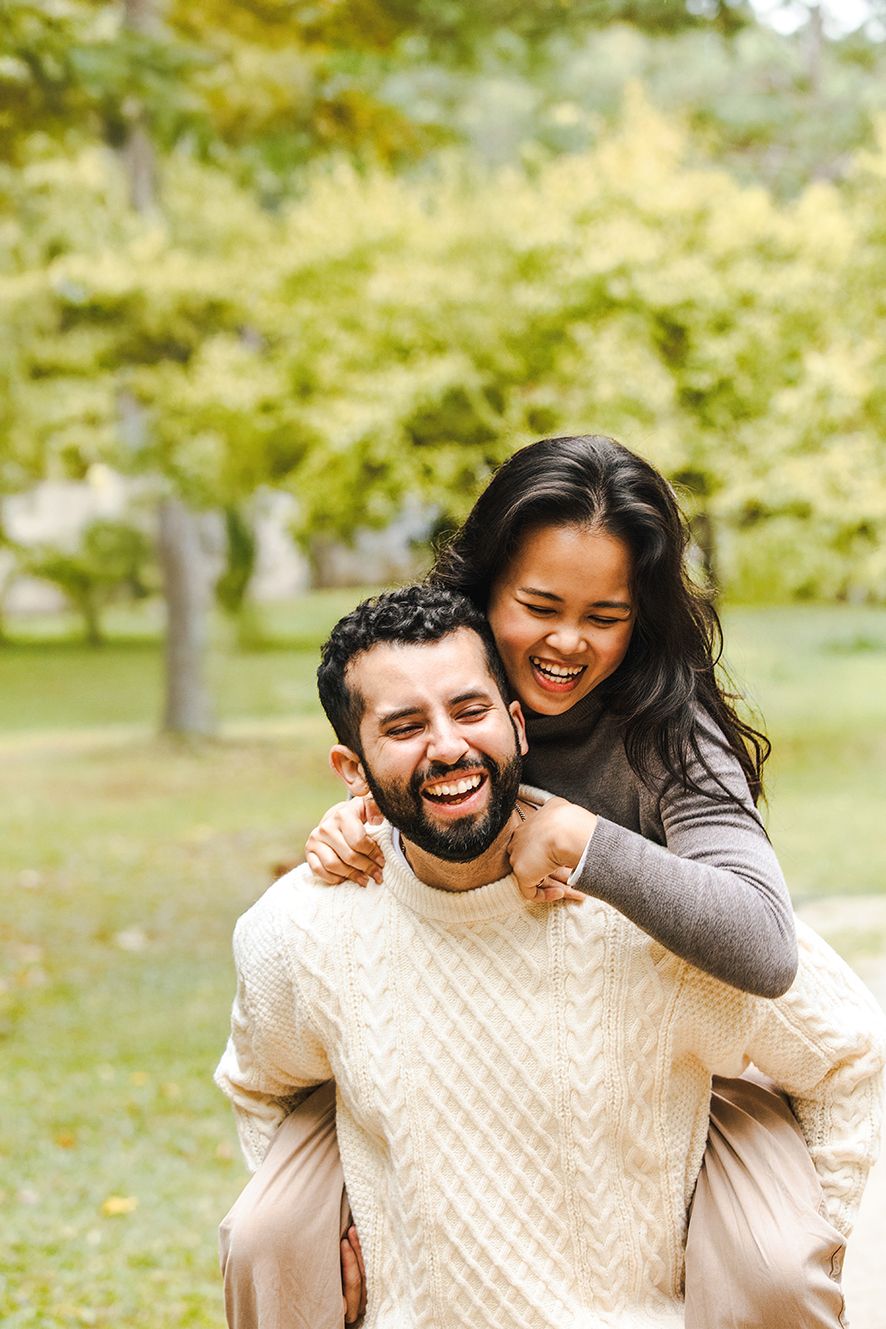 A joyful couple laughs together in an autumn park setting, with one giving a piggyback ride to the other.