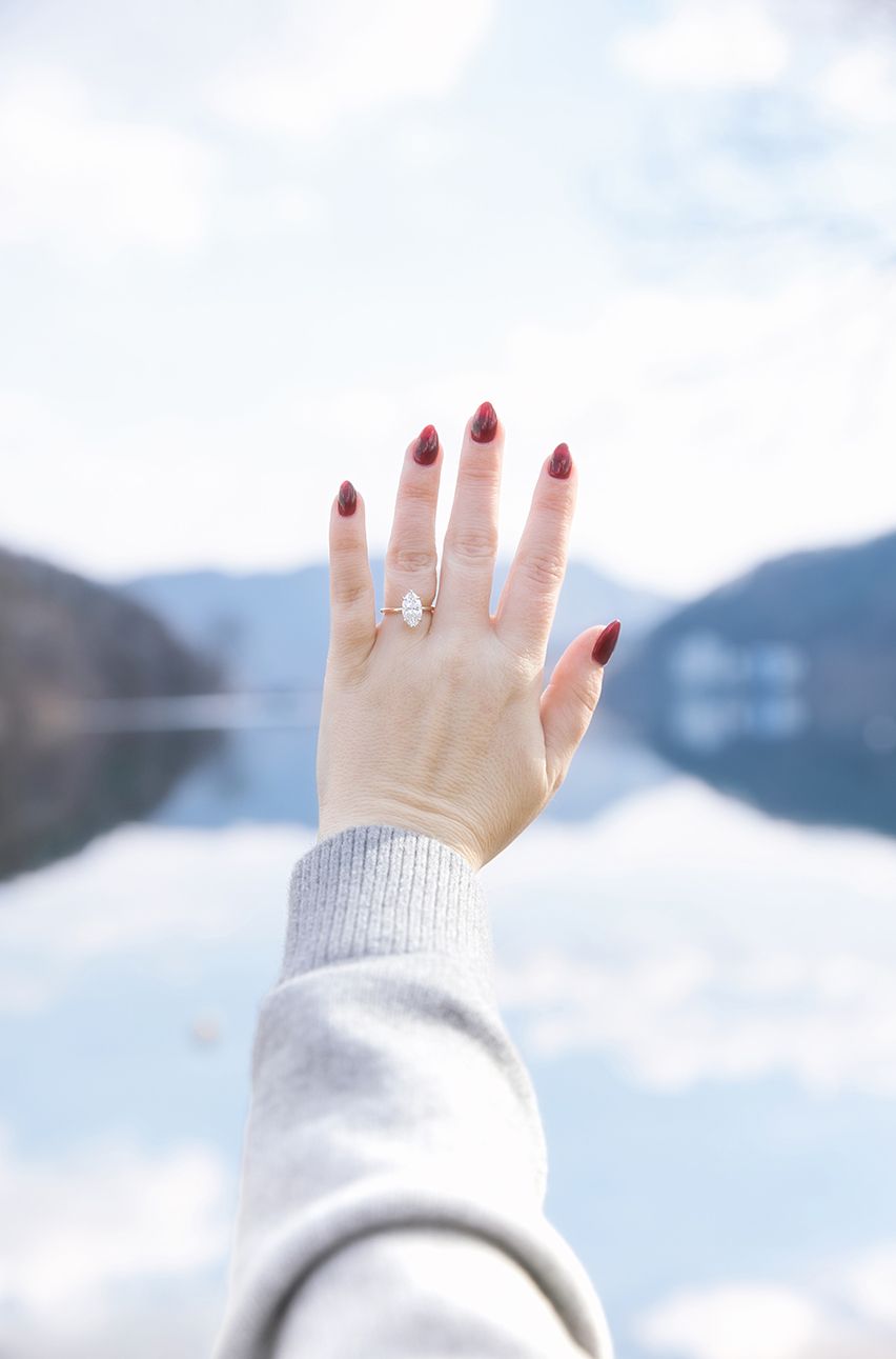 A hand with red nail polish and an engagement ring is raised against a blurry mountain lake background.