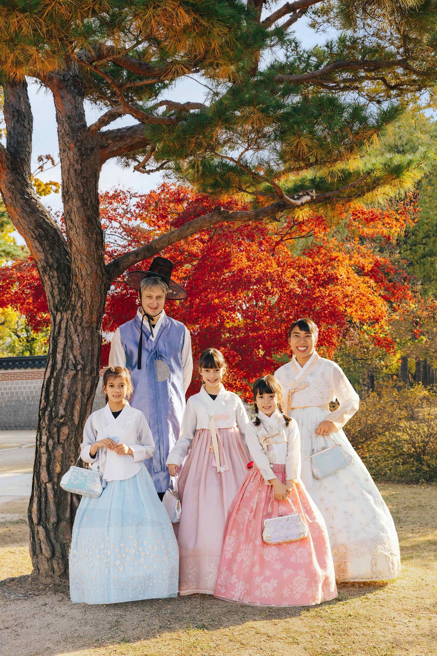 Family of five wearing traditional Korean hanbok attire posing under a pine tree with vibrant red autumn foliage.