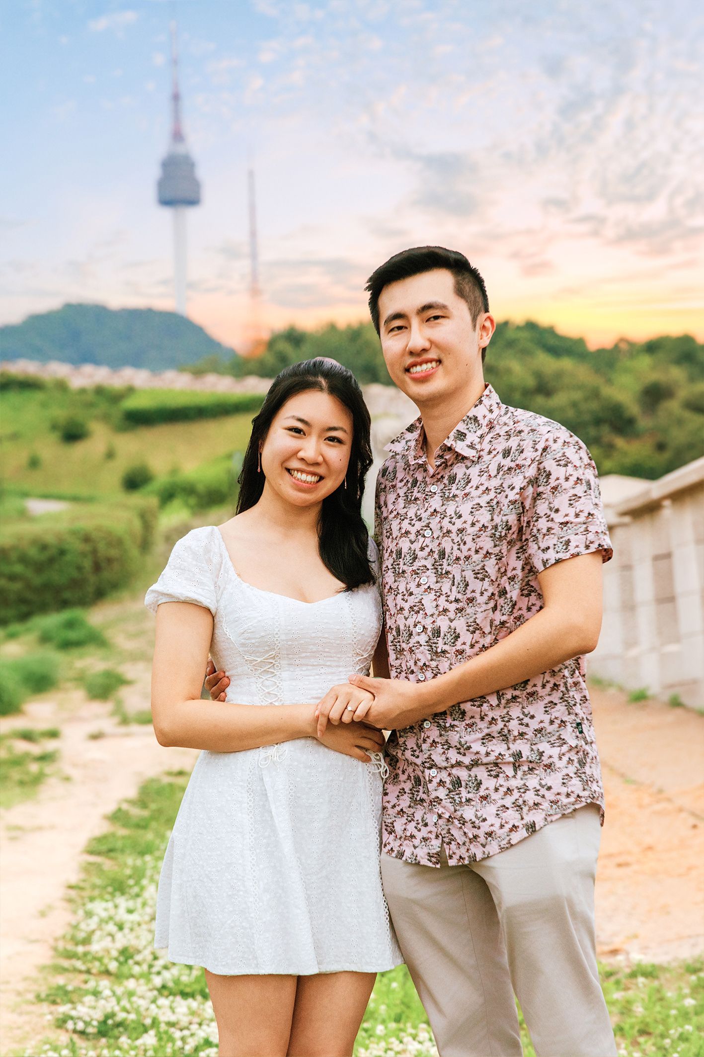 Happy Asian couple posing together outdoors near N Seoul Tower at sunset, woman in white dress, man in floral shirt.
