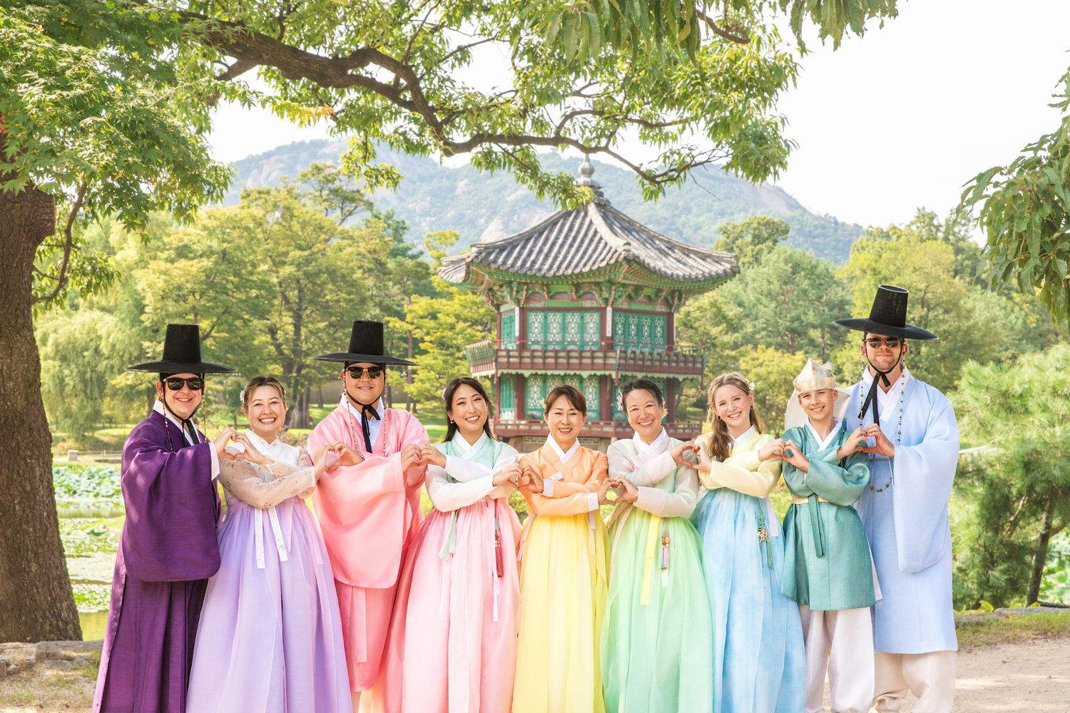 A row of people wearing colorful traditional Korean hanbok dresses stand together in a garden with pagodas and trees.