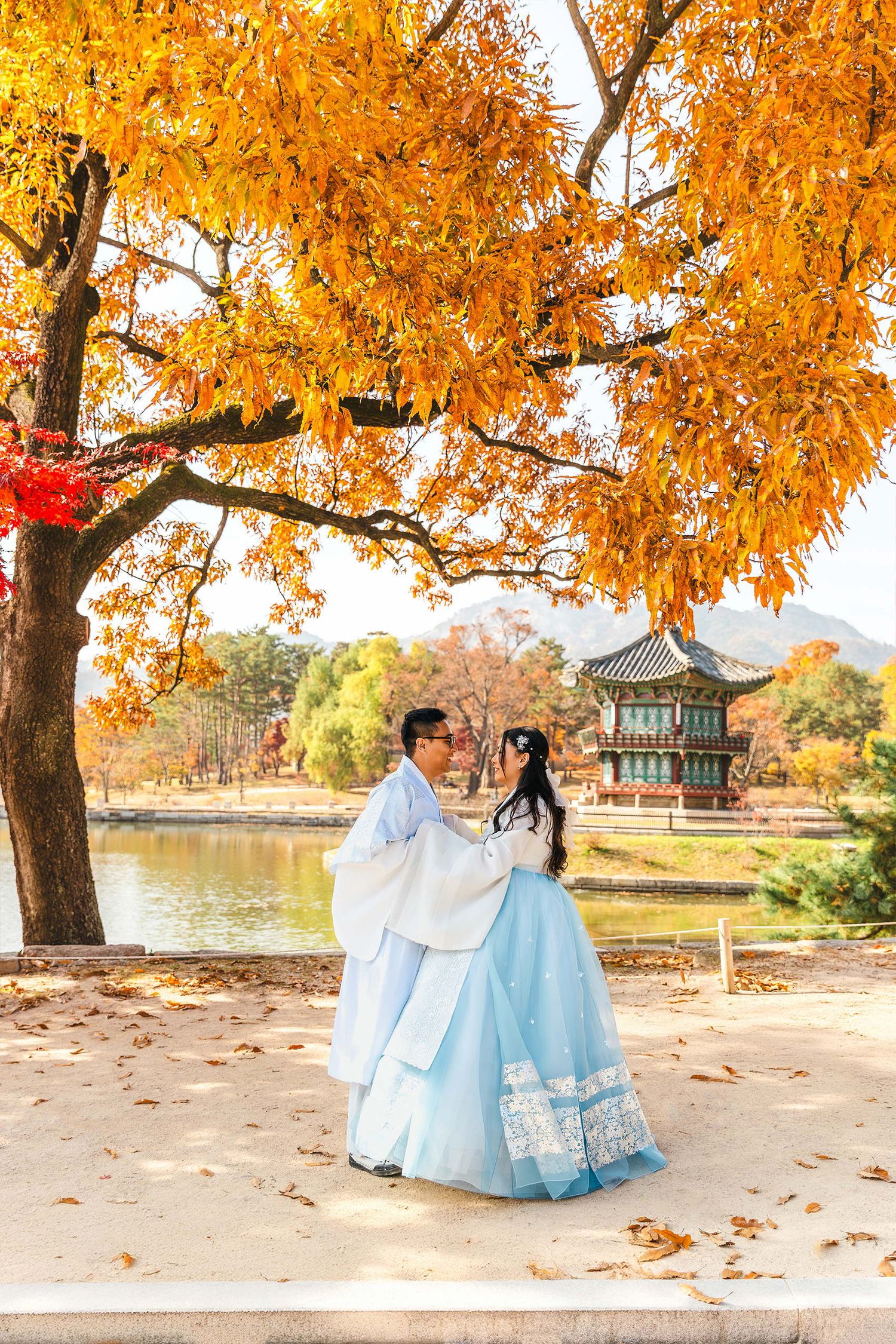 Couple in traditional Korean hanbok attire embracing under golden autumn trees near a pond at Gyeongbokgung Palace.