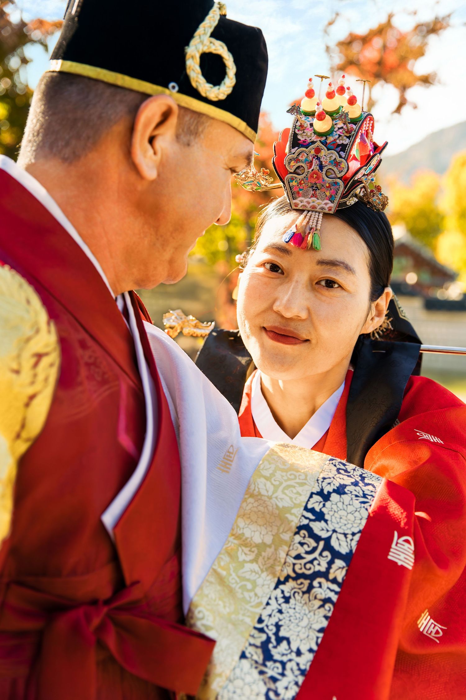 Couple in traditional Korean hanbok attire, with ornate headdress, sharing a moment outdoors in autumn.
