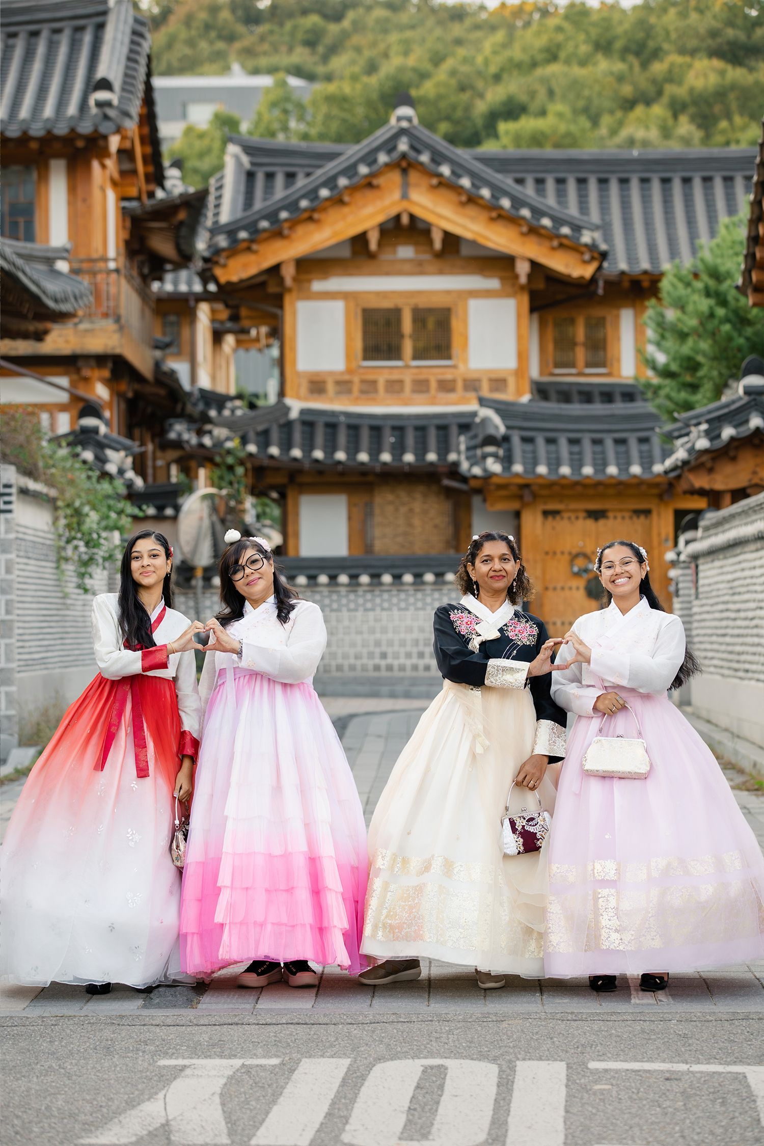 Four people wearing traditional Korean hanbok dresses pose in front of traditional Korean architecture with curved tile roofs.