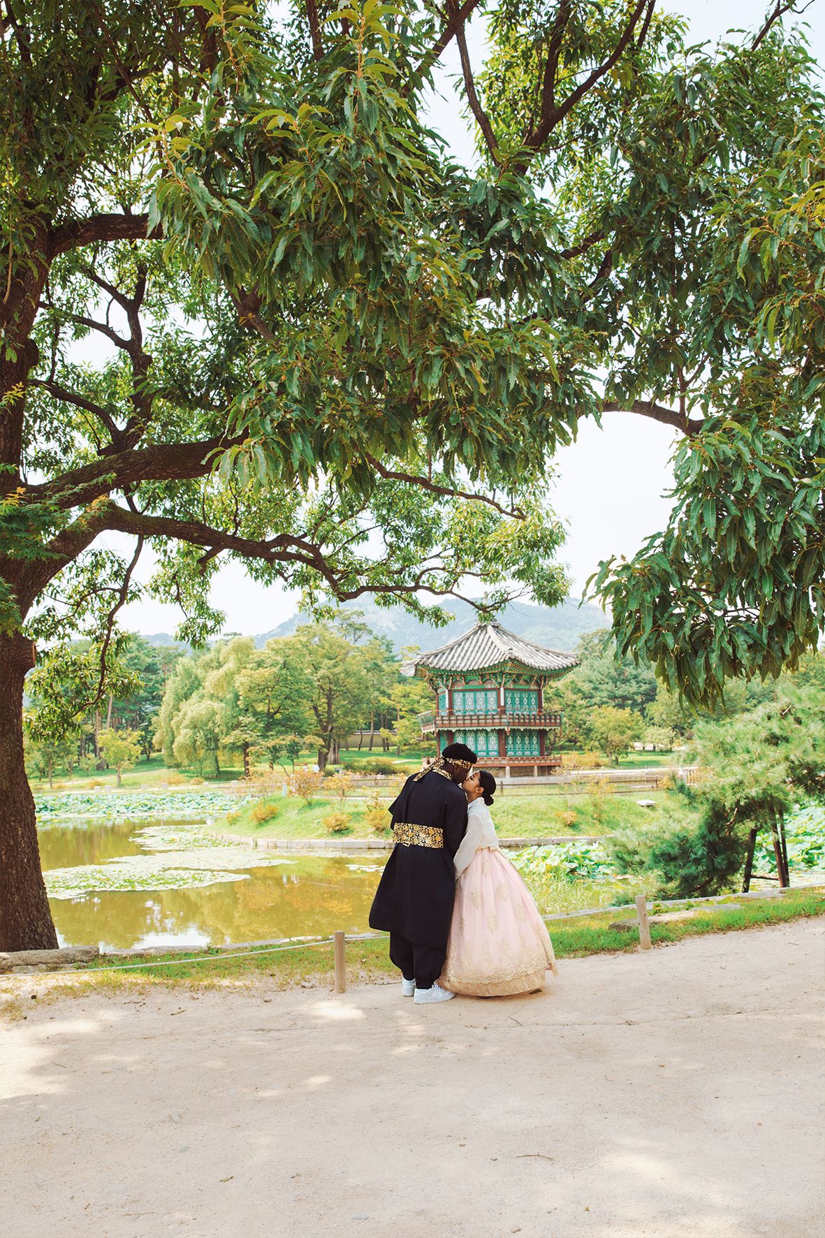 A traditional Korean pavilion stands near a pond surrounded by lush trees in a tranquil garden setting.