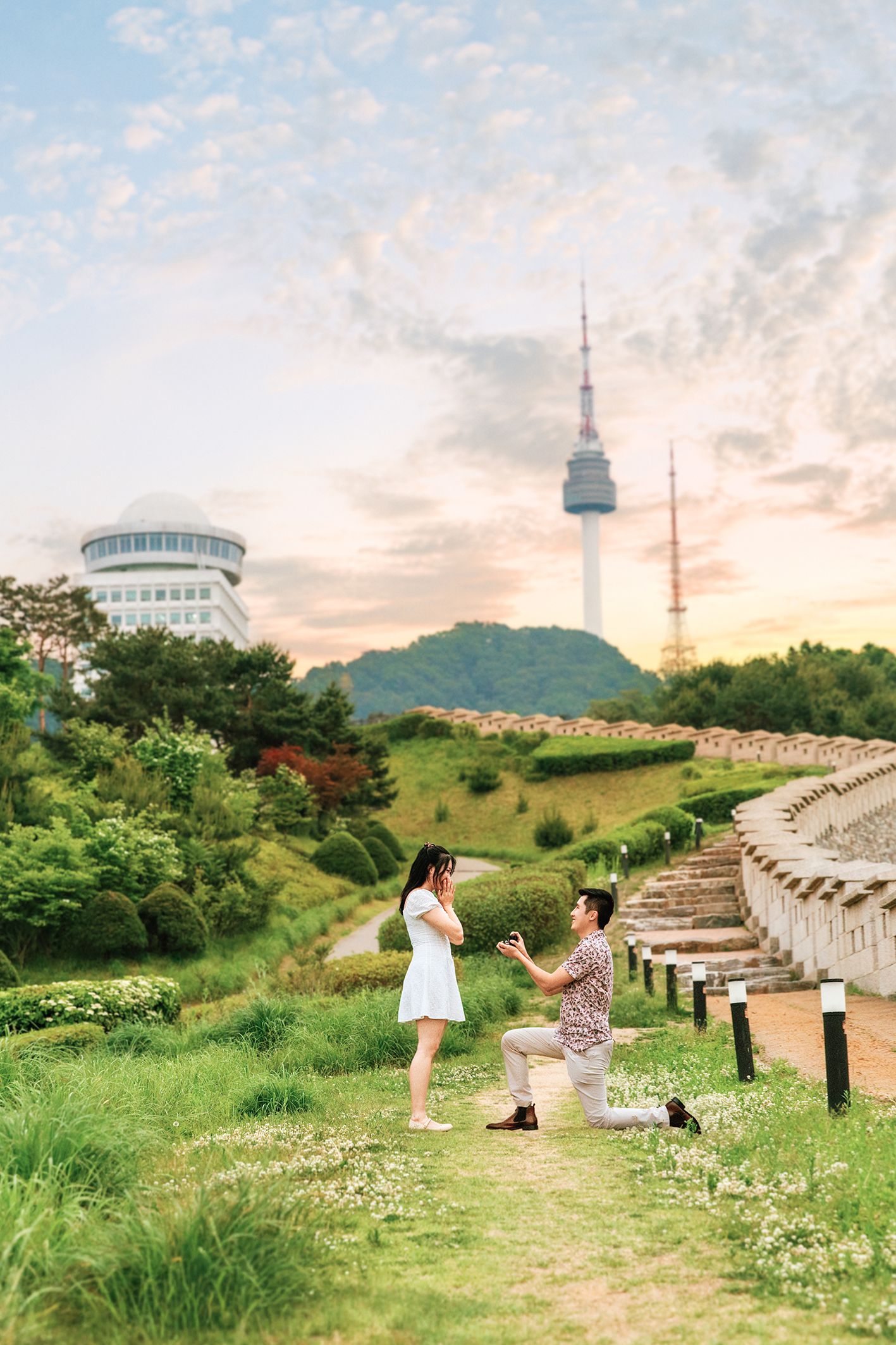 Man proposes to woman on one knee in a lush garden with N Seoul Tower and scenic hilltop in the background.