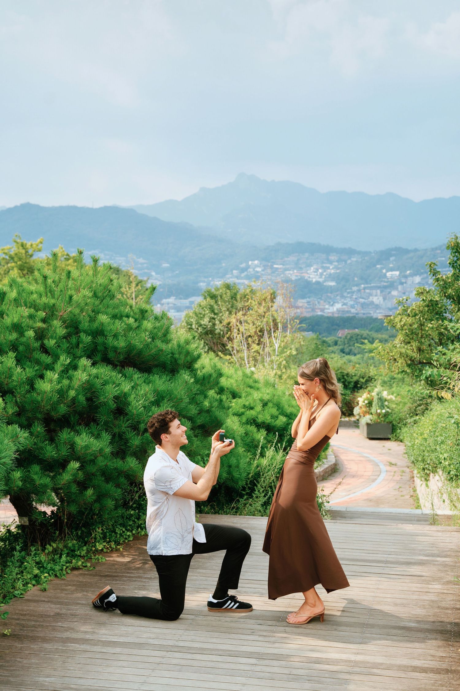 Man proposes on one knee to woman on scenic overlook with lush greenery and mountain views in background.