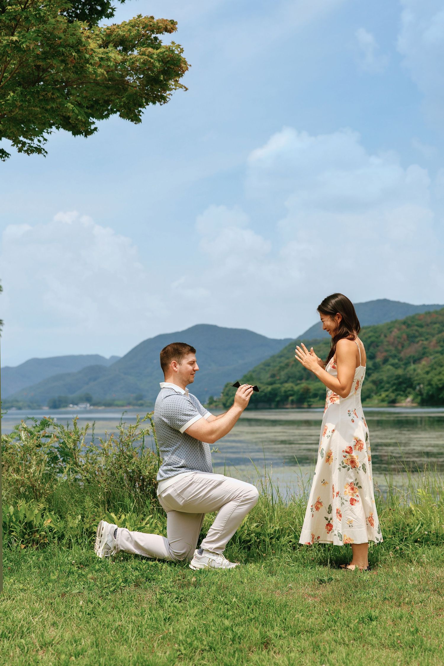 Man proposes on one knee to woman in floral dress by scenic lake with mountains in background on sunny day.