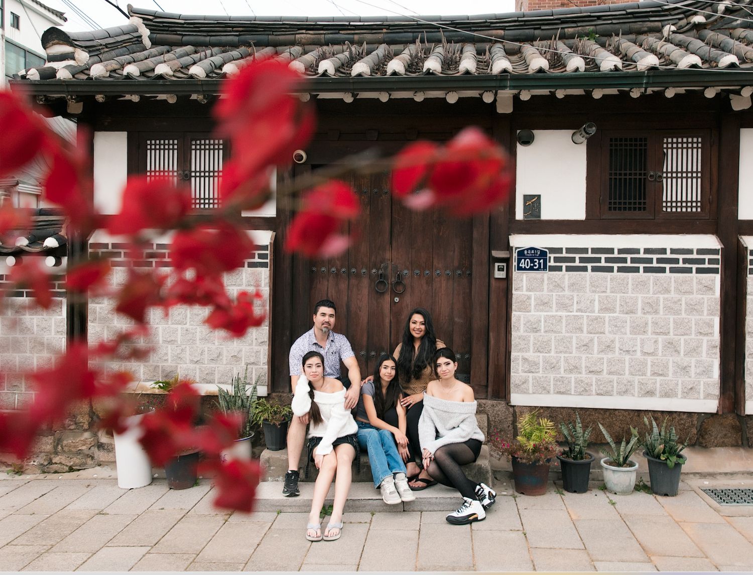 Group of friends posing in front of a traditional Korean hanok building with red flowers in foreground.