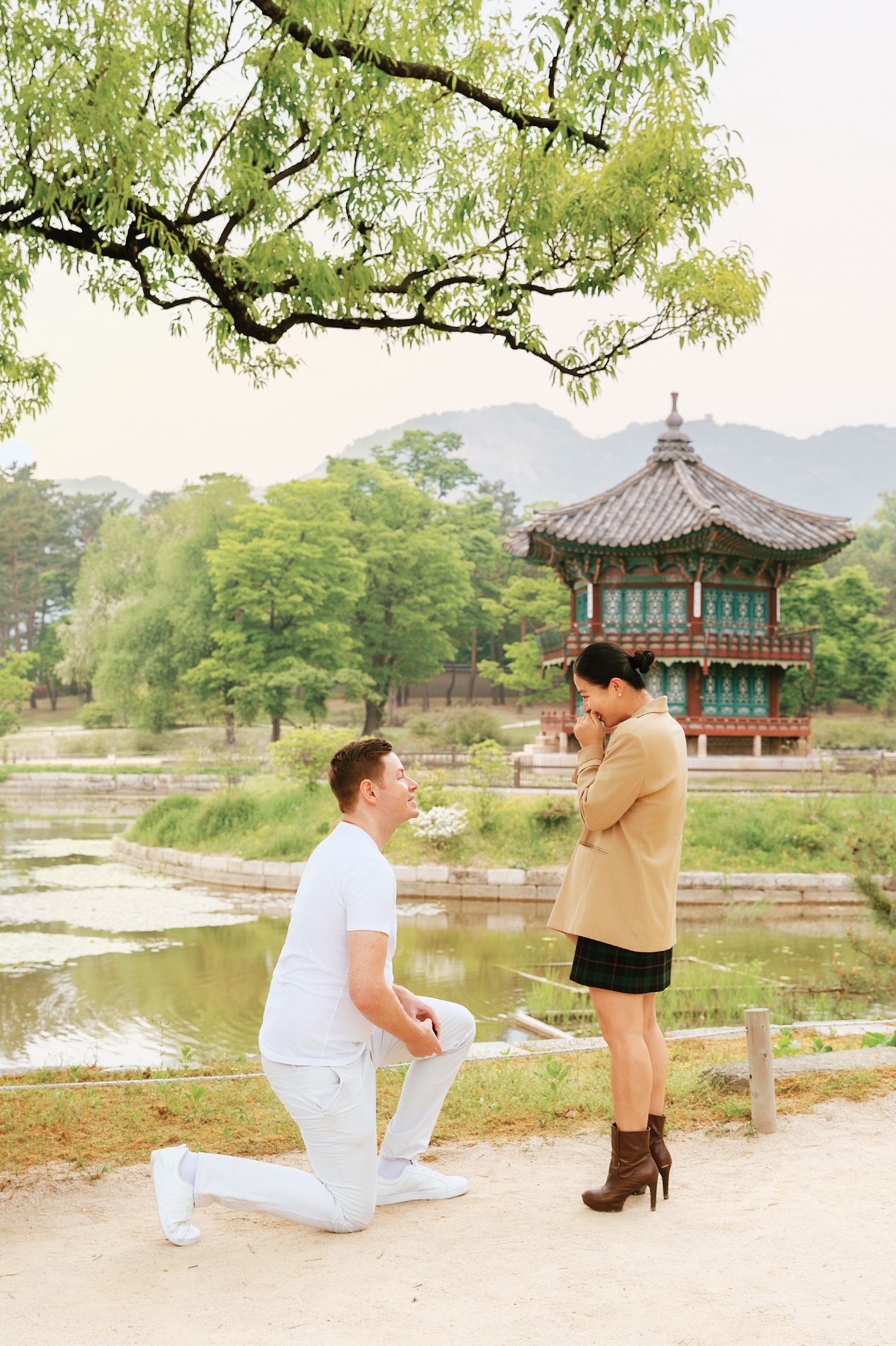 Man proposing on one knee to woman near a traditional Korean pavilion by a serene pond with lush green trees.
