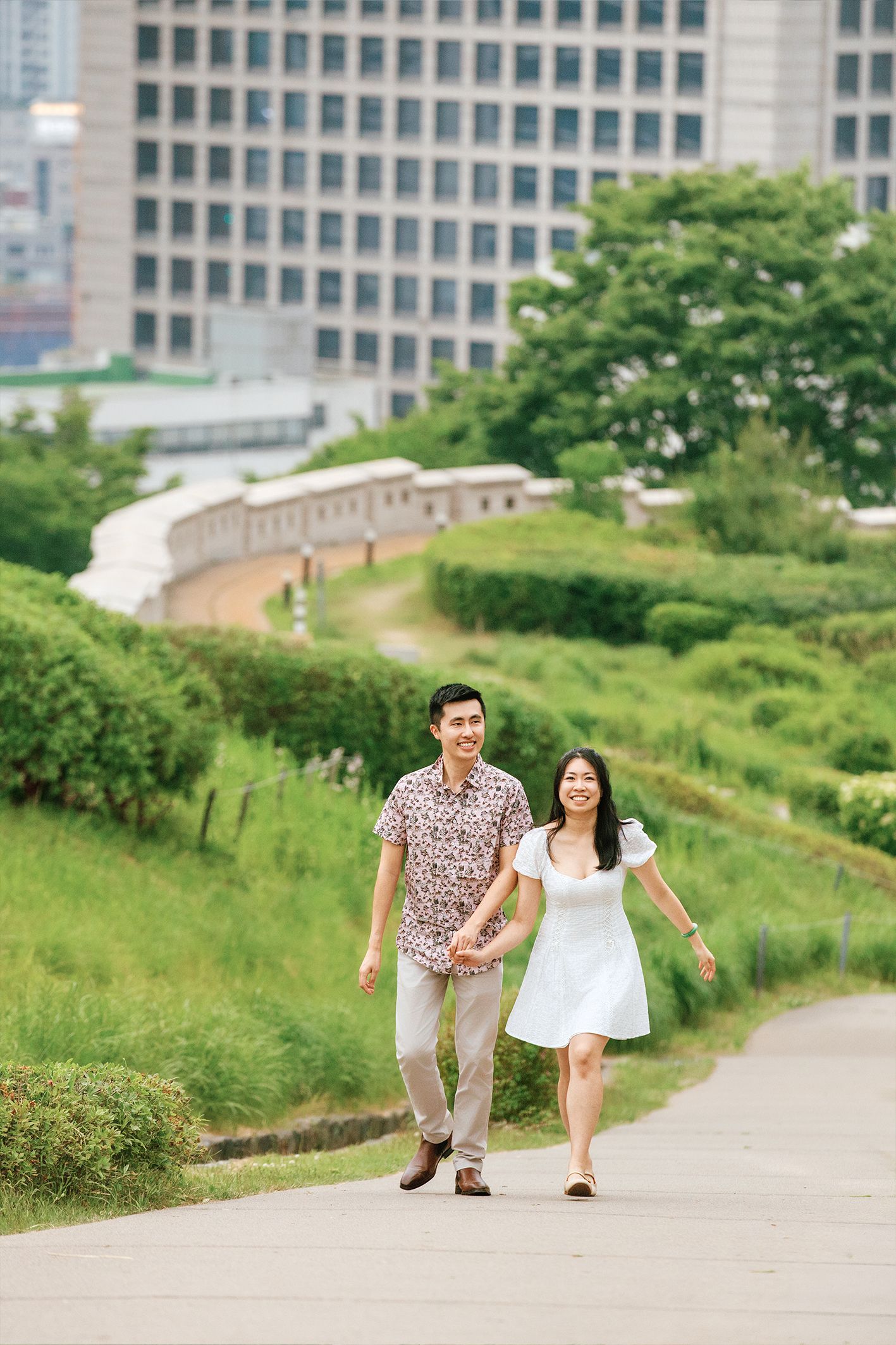 A smiling Asian couple walks hand-in-hand on a path surrounded by lush greenery with city buildings in the background.