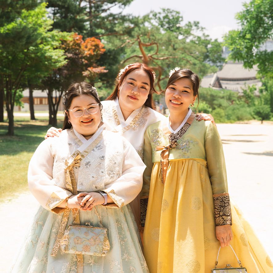 A group in traditional Korean hanbok dresses pose together in a bright outdoor park setting filled with greenery.