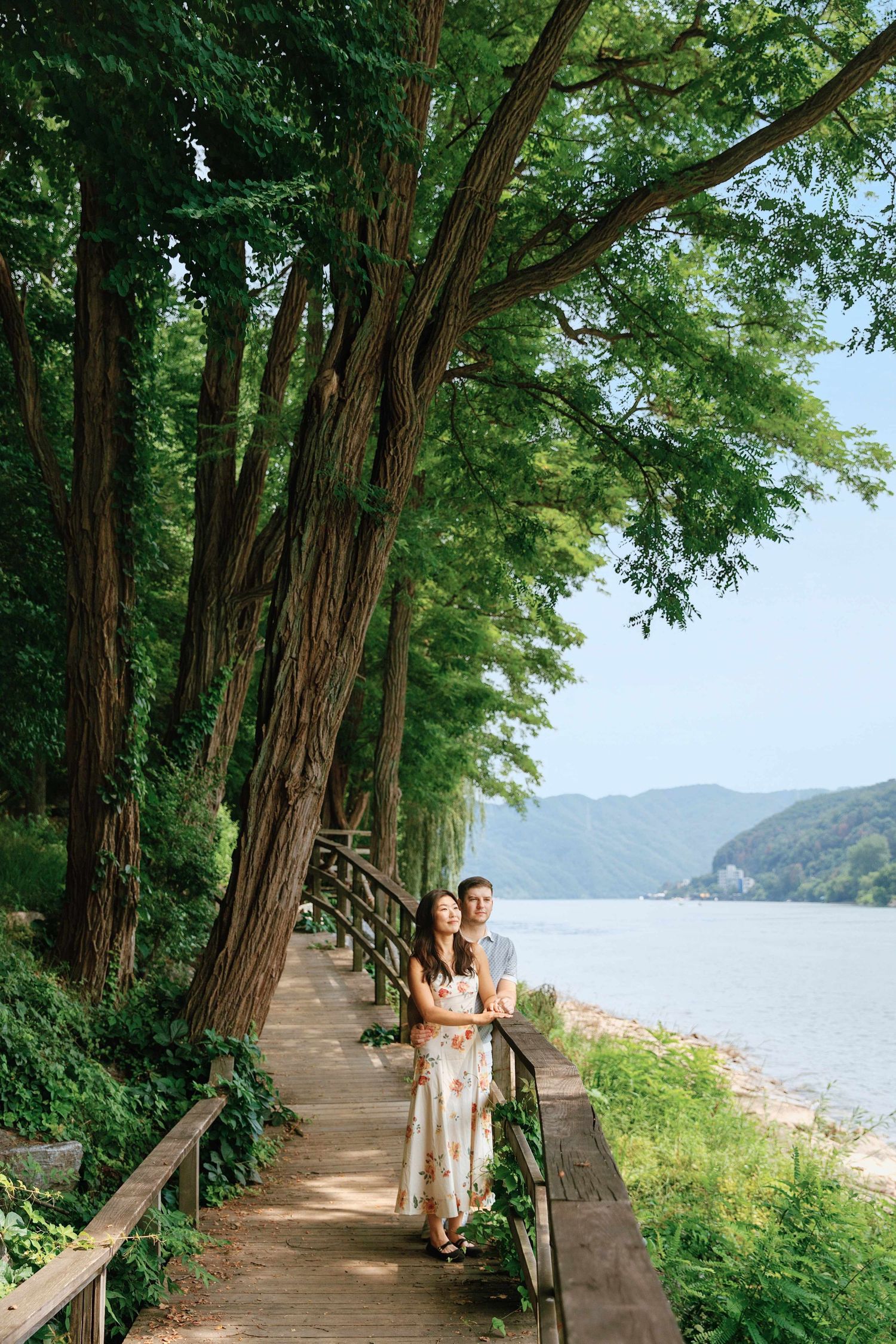 Couple embracing on a wooden boardwalk path lined with tall green trees beside a calm lake with mountain views.