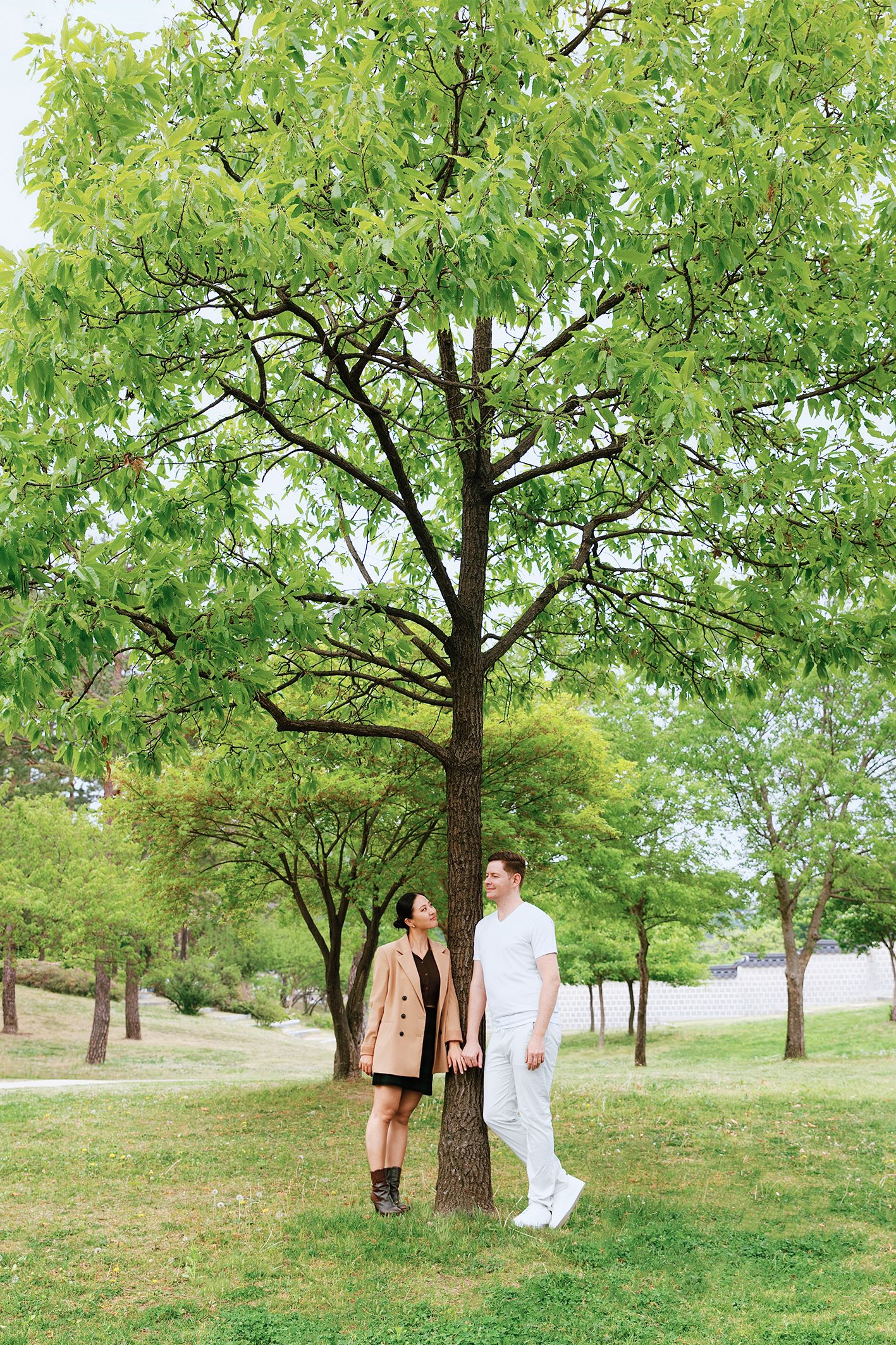 Couple holding hands under a large green tree in a lush park, woman in beige coat, man in white outfit.