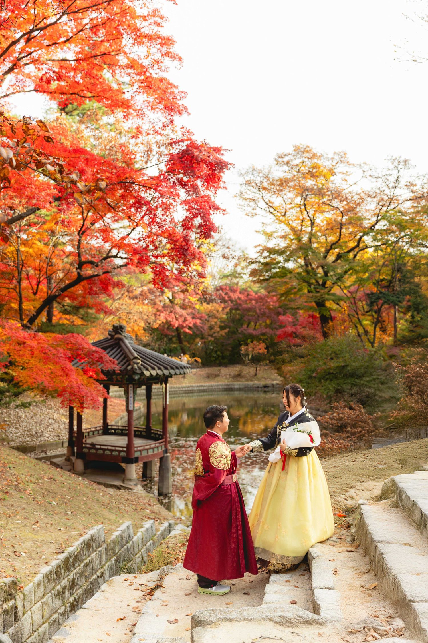 Two women in traditional Korean hanbok attire stand by a pond surrounded by vibrant autumn foliage in a historic garden.