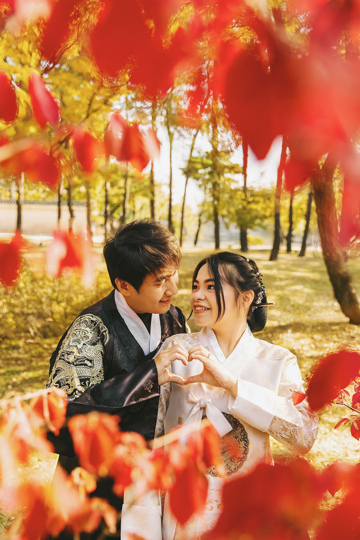 Couple in traditional Korean hanbok making heart shape with hands among vibrant red autumn leaves in a park.