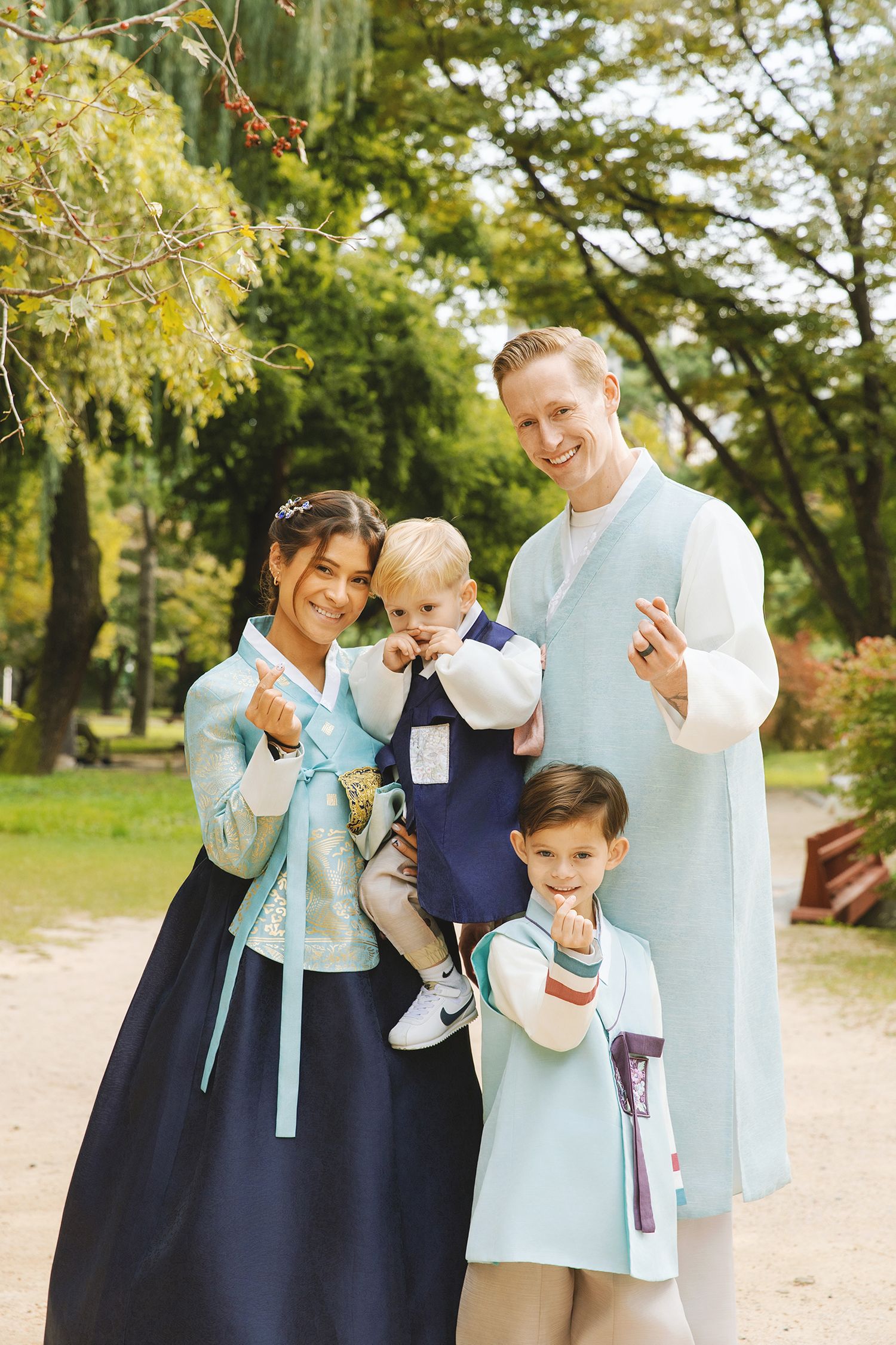 A family shares joyful moments together on a dirt path surrounded by lush green trees in a park setting.