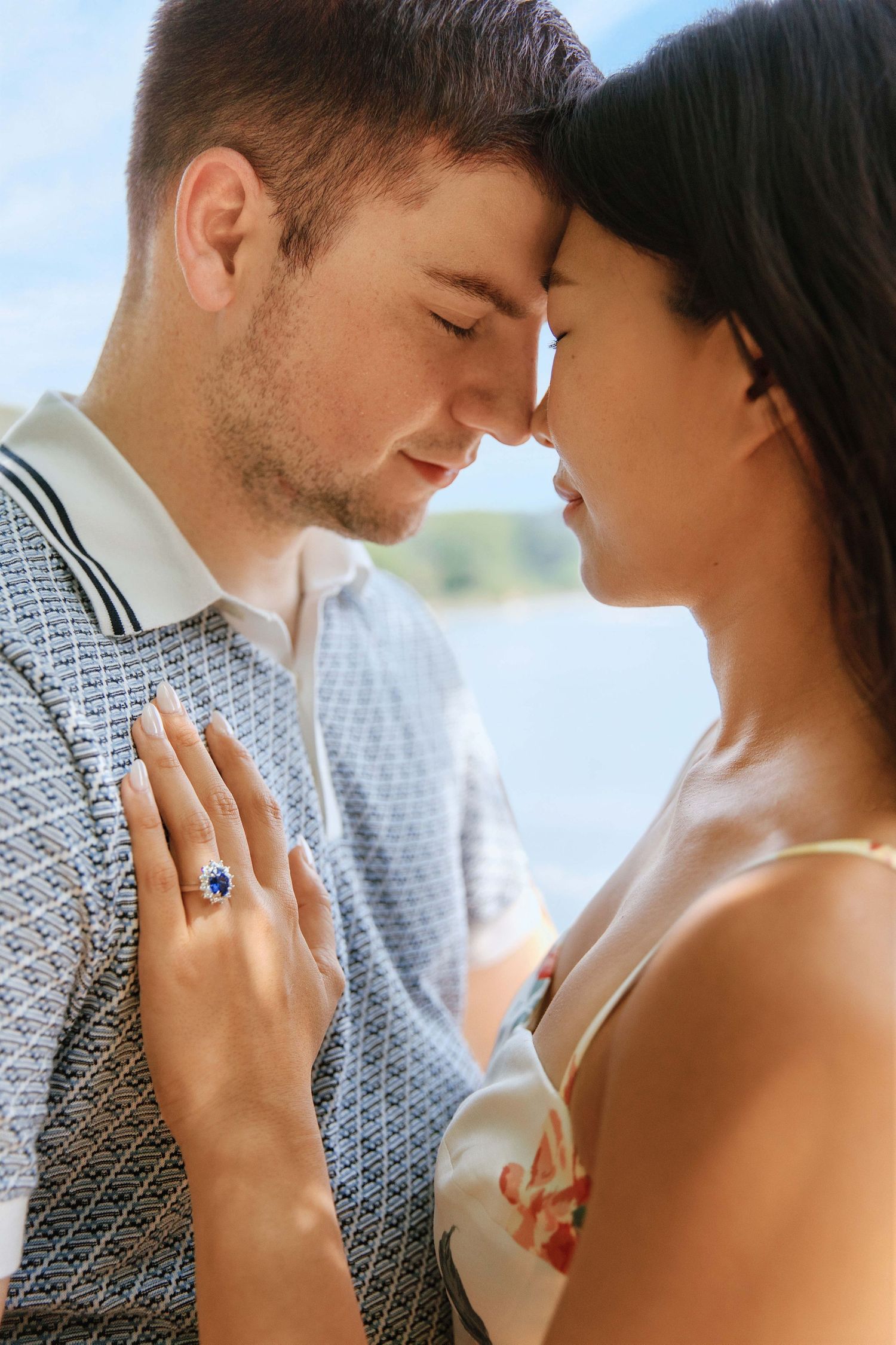 Romantic couple touching foreheads outdoors, woman displaying a stunning sapphire and diamond engagement ring on her hand.