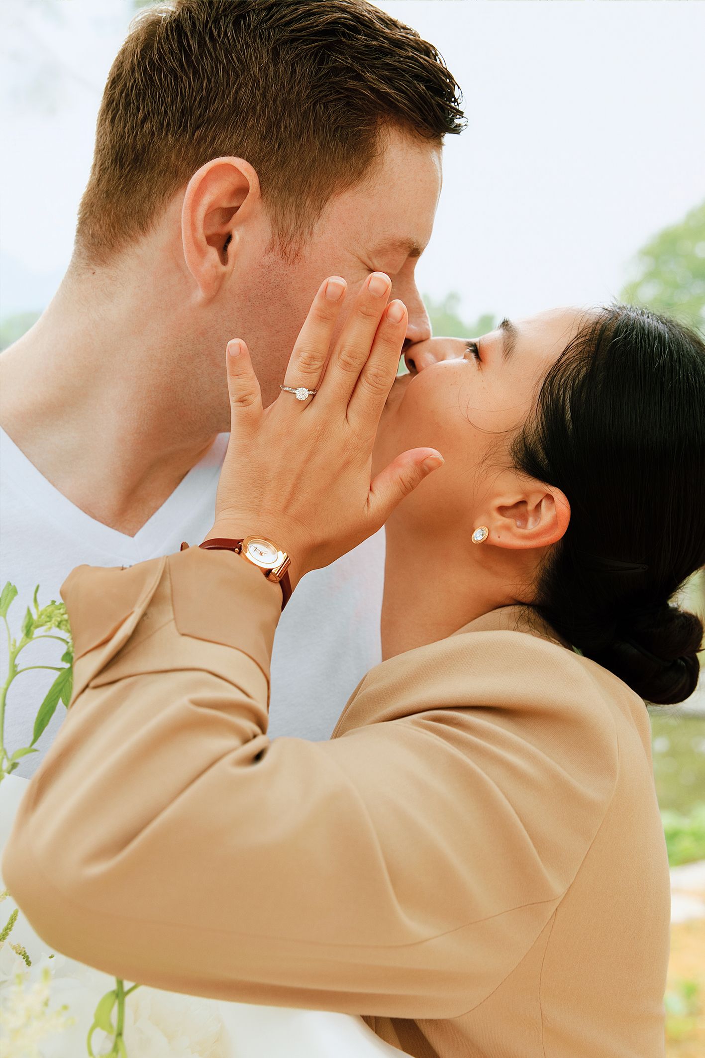 Couple sharing a romantic kiss, woman wearing engagement ring and tan blazer with a gold watch.
